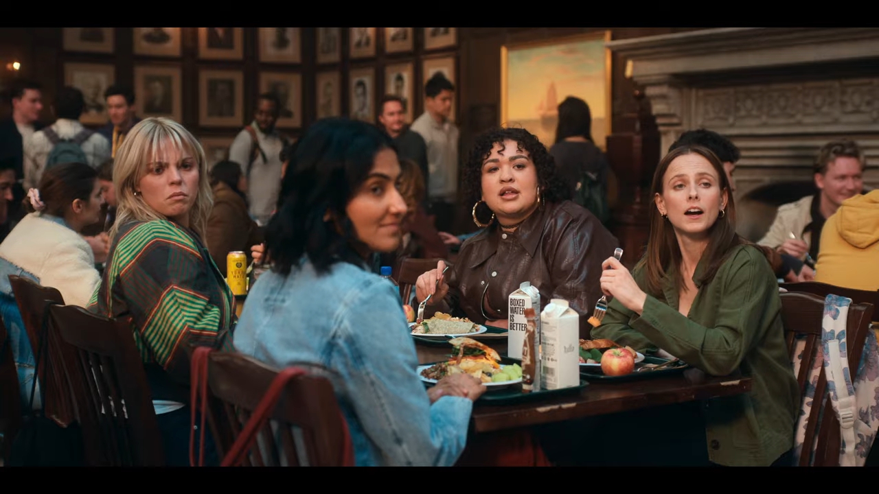 Four female college students sit at a crowded cafeteria table, looking surprised. From the show "Sex Lives of College Girls"