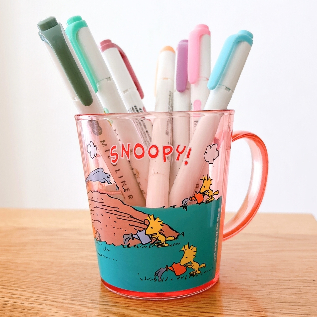 スヌーピーのイラストが描かれたマグカップに、色とりどりのペンが入っている。