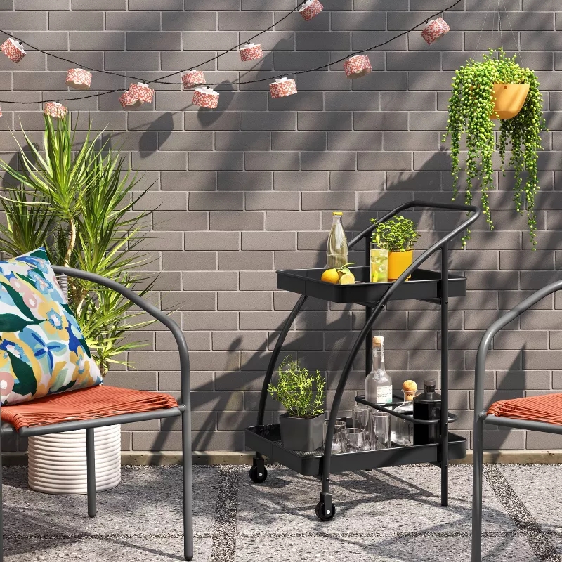 Outdoor patio with a black bar cart holding drinks and plants, surrounded by metal chairs; brick wall adorned with small string lights