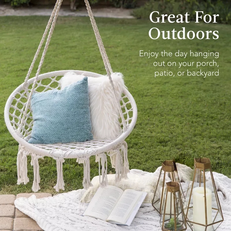 A cozy outdoor setup with a macramé swing chair, two pillows, an open book, and decorative lanterns on a patio, promoting relaxation