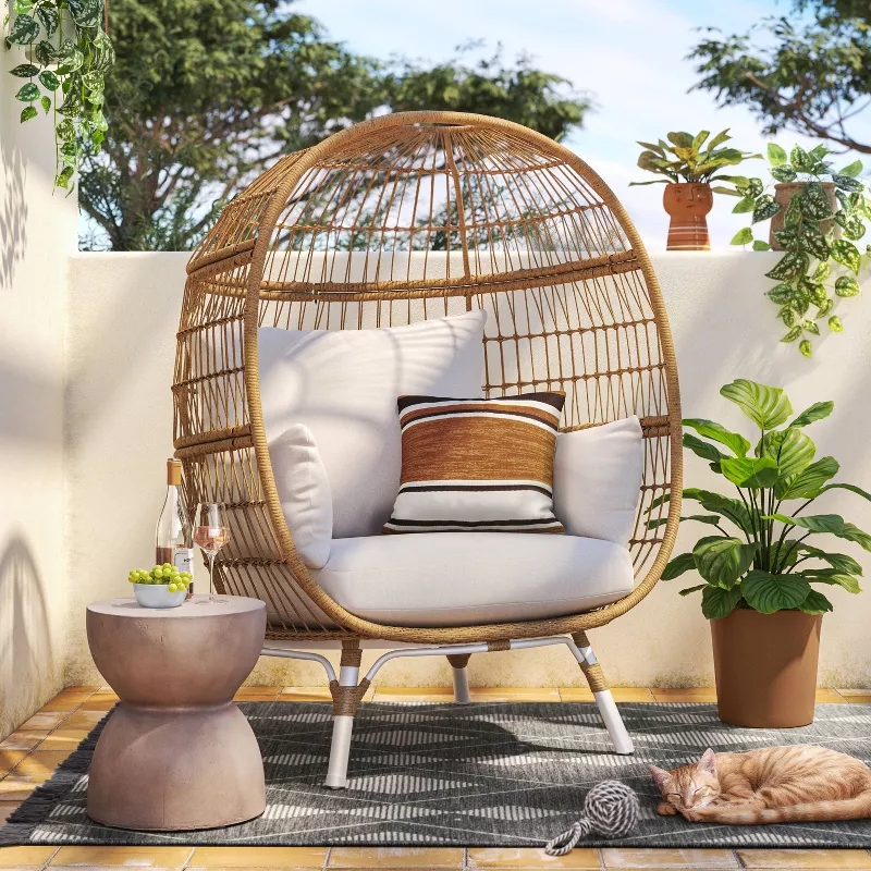 Egg-shaped wicker chair with cushions on a patio. Nearby, a small table holds drinks and snacks. A cat sleeps on a rug beside a potted plant