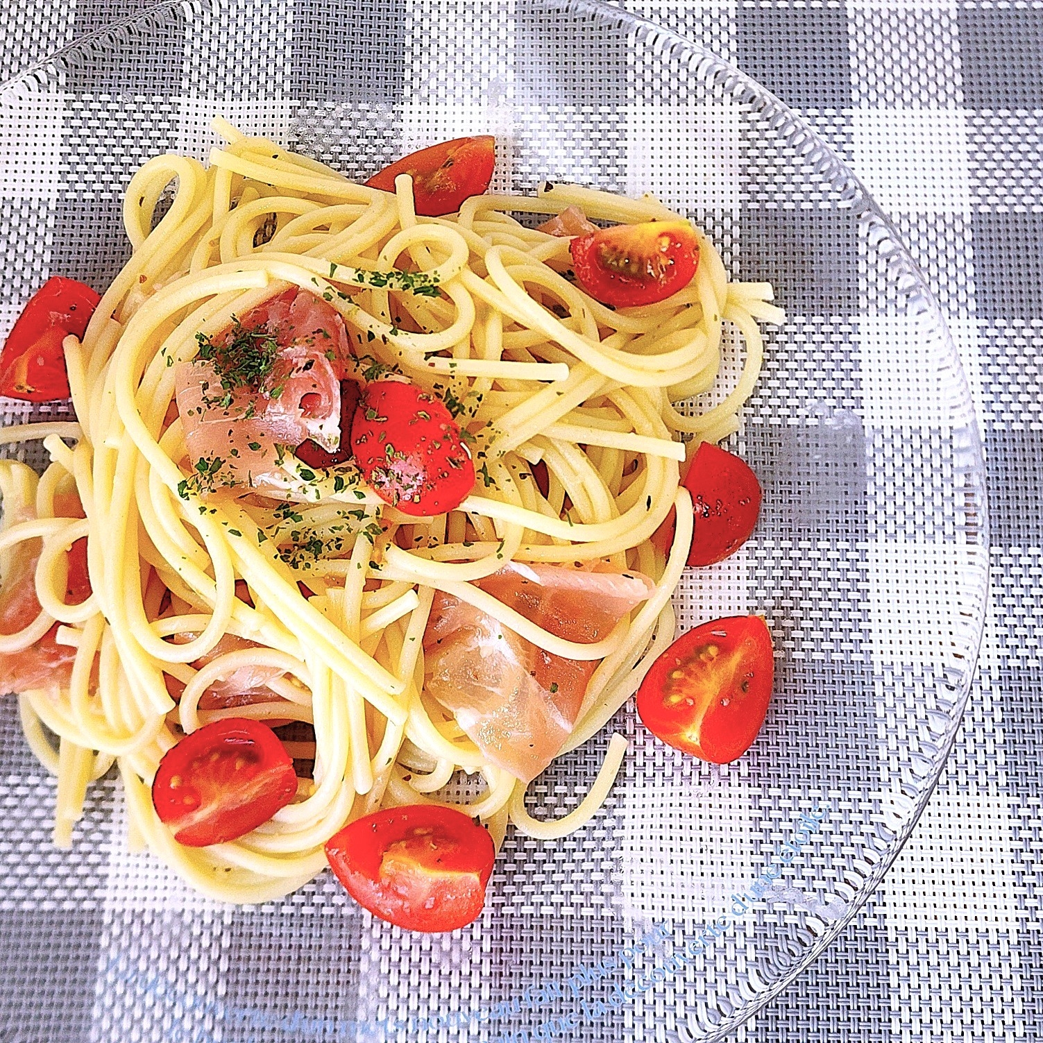 テーブル上の透明な皿に盛られたスパゲッティ。トマトとハムがのっている。