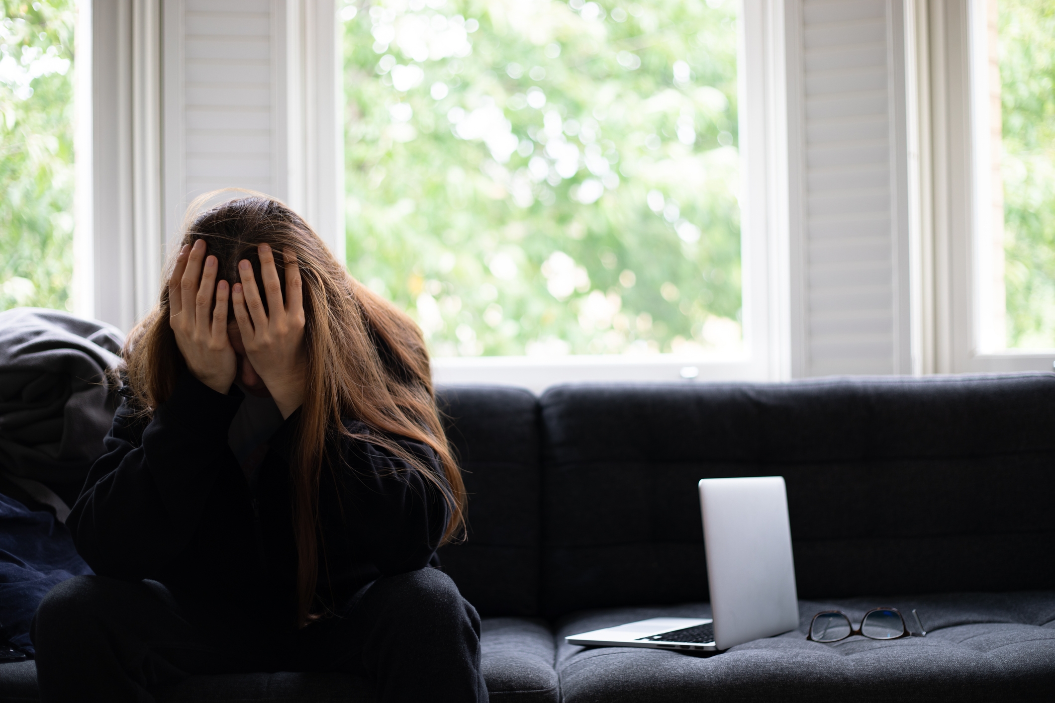 A person sits on a couch with their head in their hands beside a laptop and glasses, suggesting stress or overwhelm in a work context