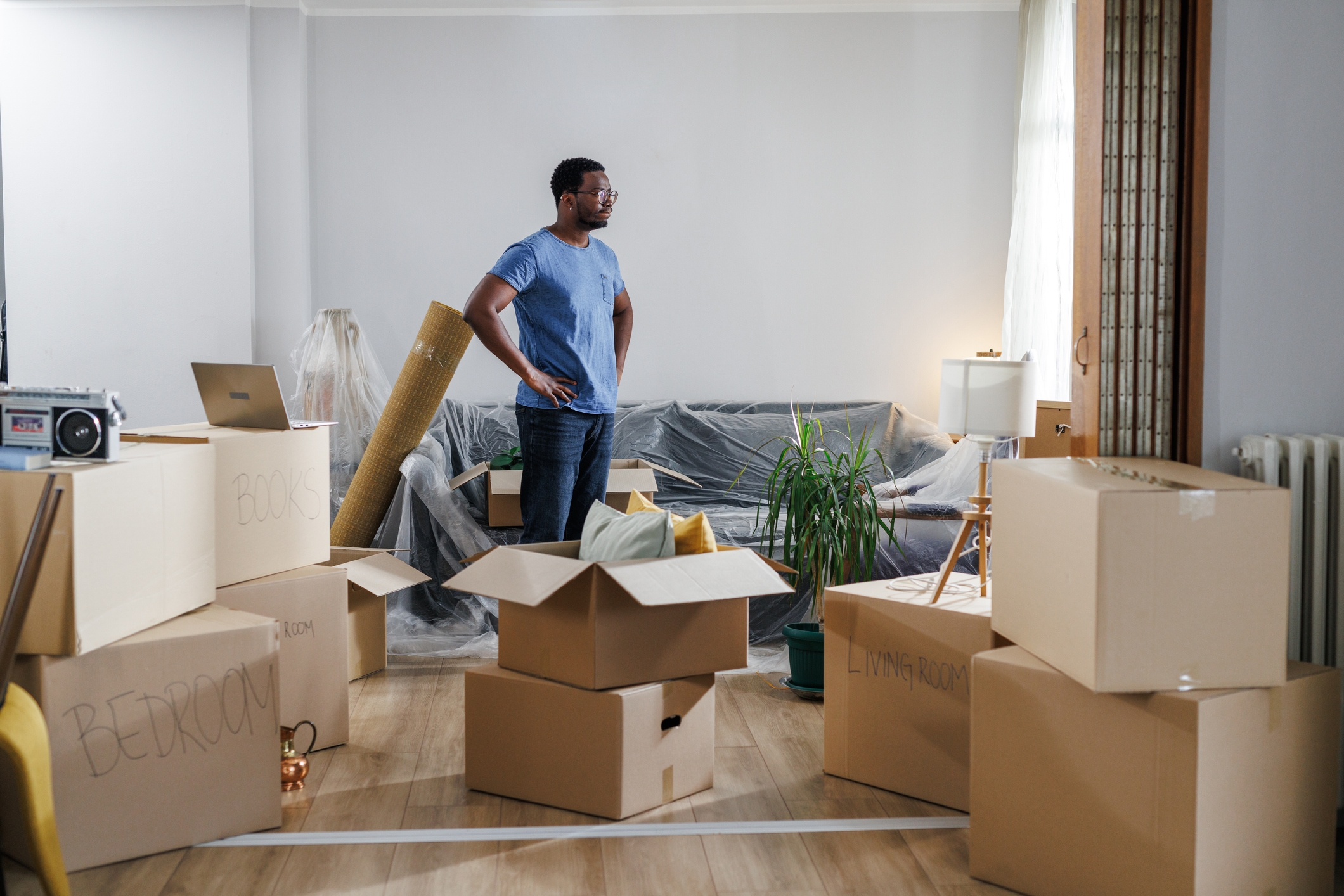 Person stands in a living room surrounded by moving boxes, looking contemplative. A laptop and unpacked items are visible