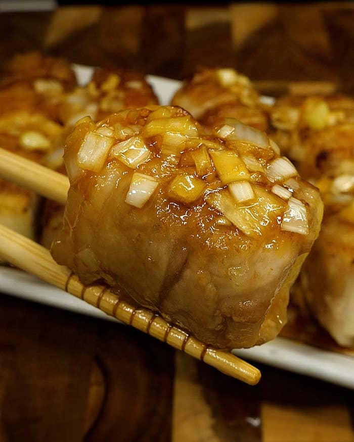 箸で持たれた一口サイズの料理。刻んだ玉ねぎとソースがかかっている。奥に複数の同様の料理が見える。