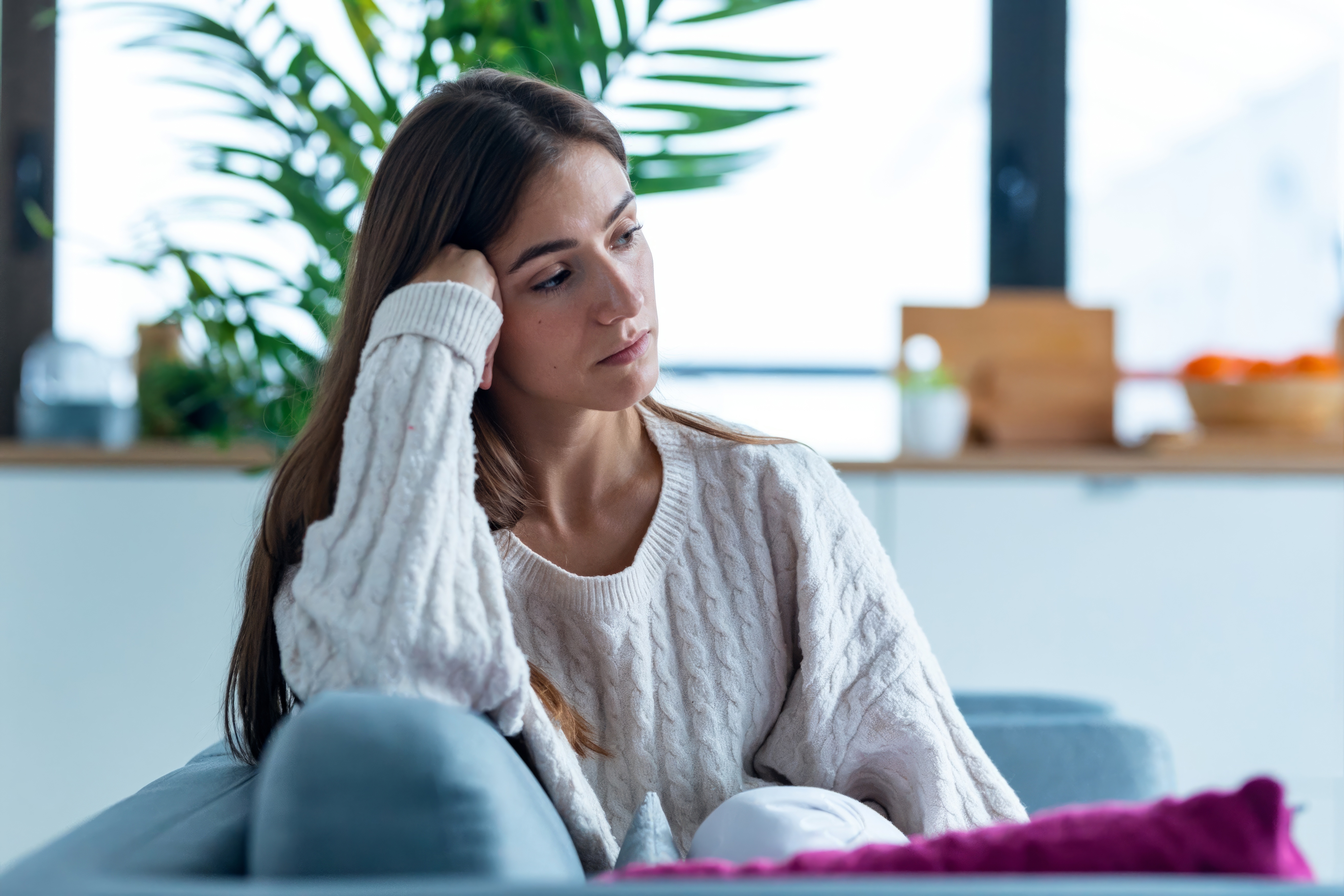 Woman sitting on a couch, deep in thought, wearing a cozy knit sweater, with plants in the background