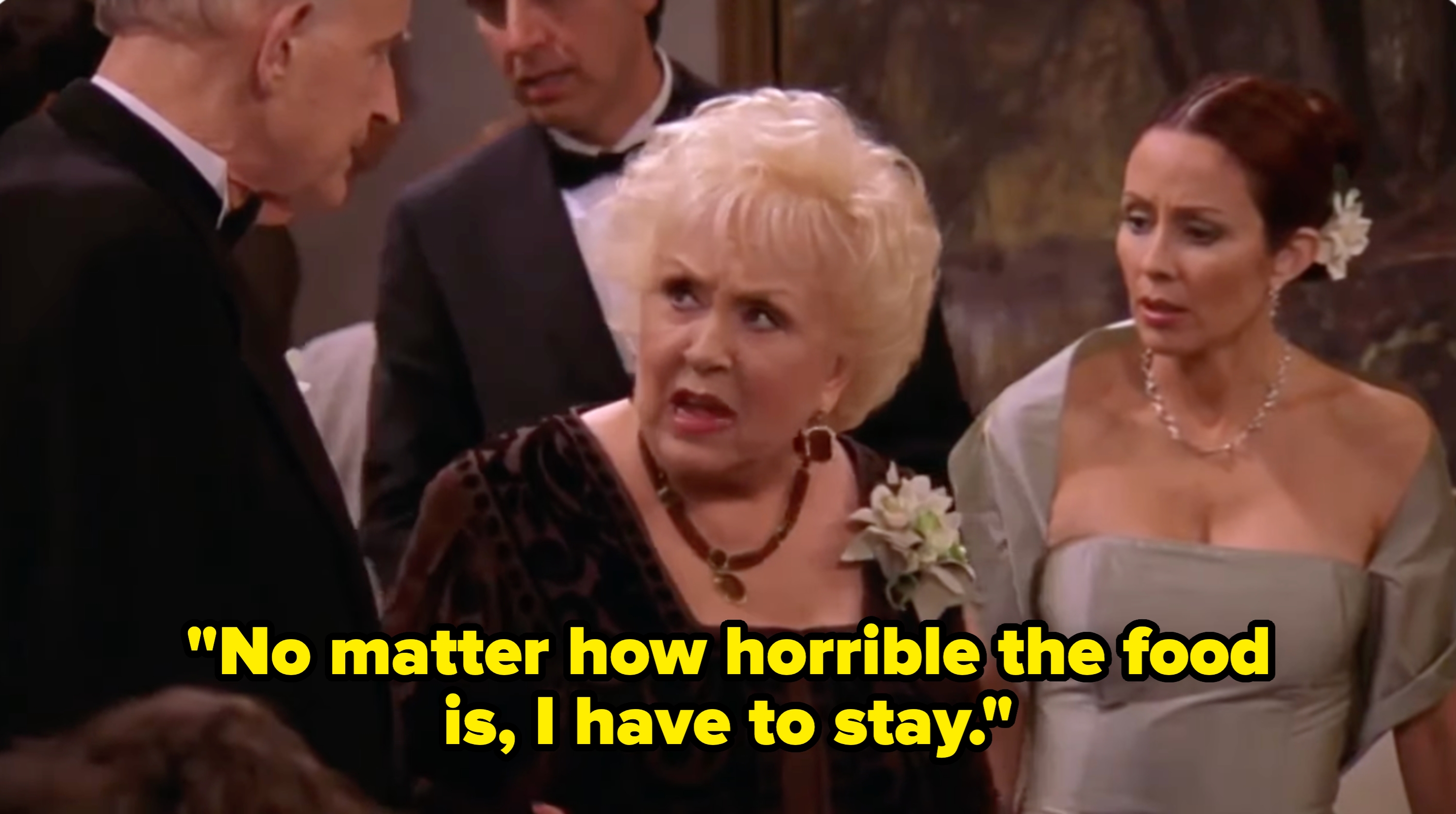 An older woman and a younger woman in formal attire look concerned at a wedding reception. Guests in background