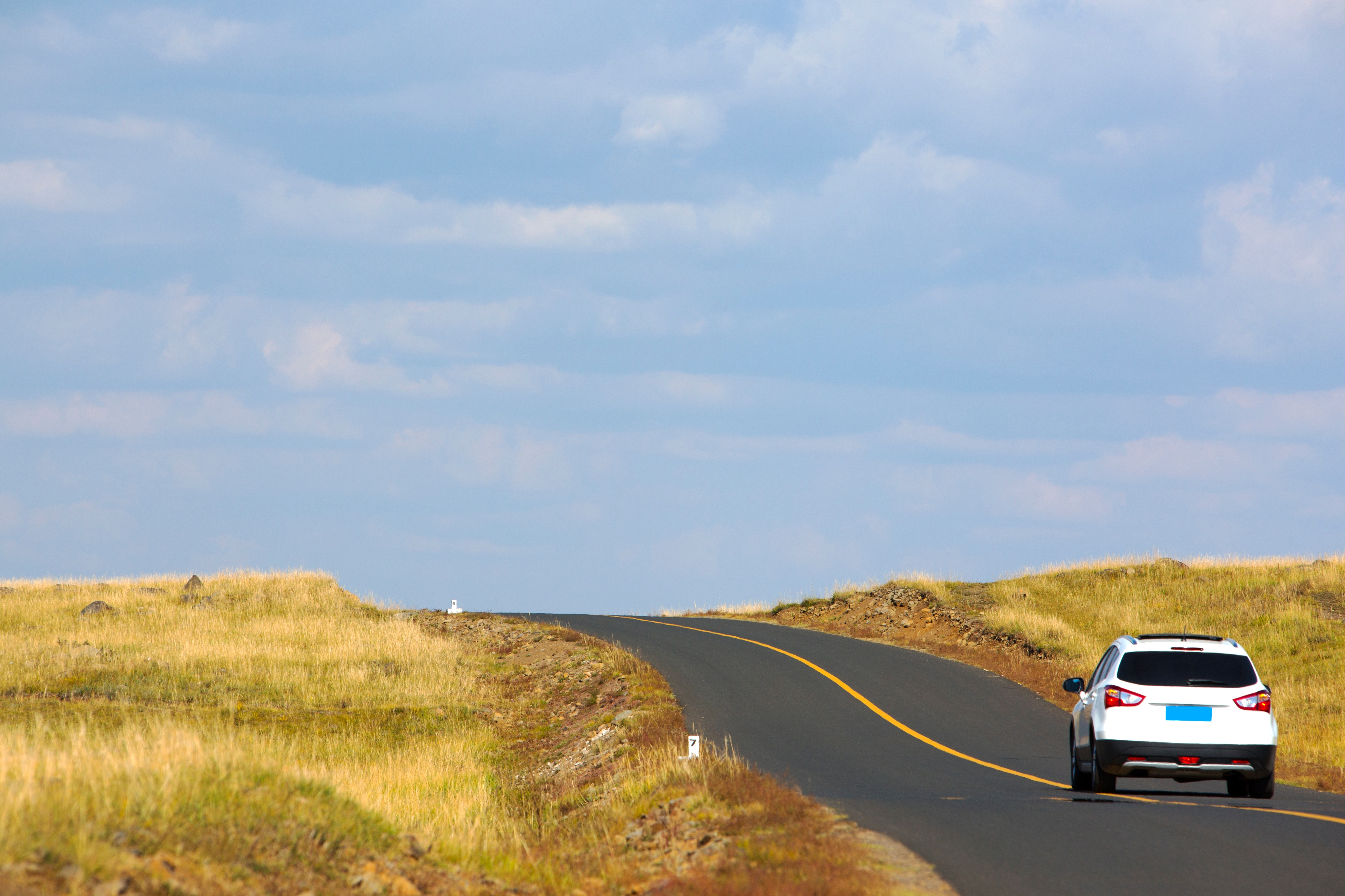 A white car drives on an empty rural road surrounded by open grassland under a cloudy sky