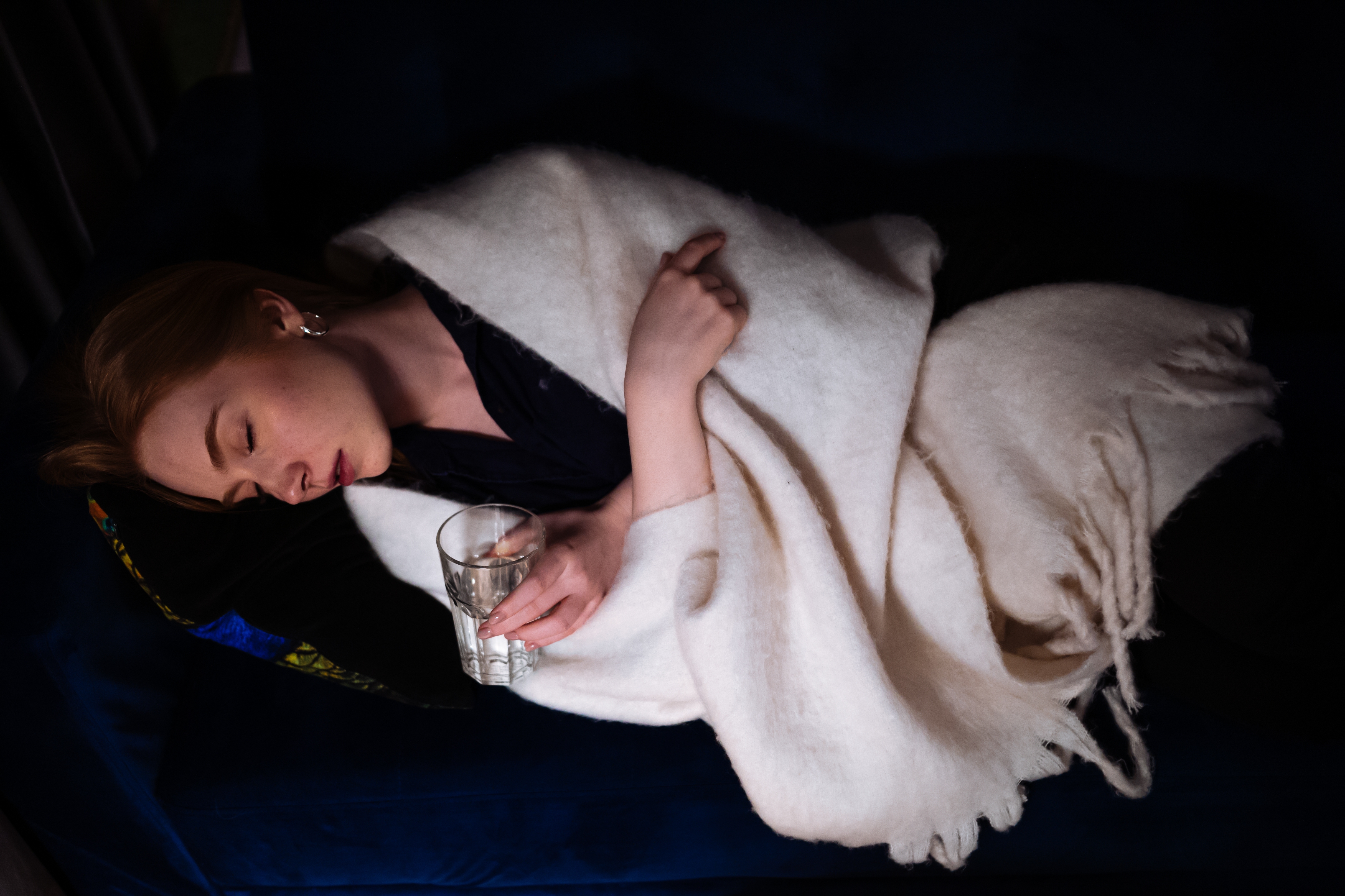 Person som sover på en mørk sofa, pakket inn i et lett teppe, holder et glass klar væske