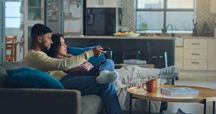 A couple sits on a couch at home, relaxed and watching TV, with a cozy living room setting in the background