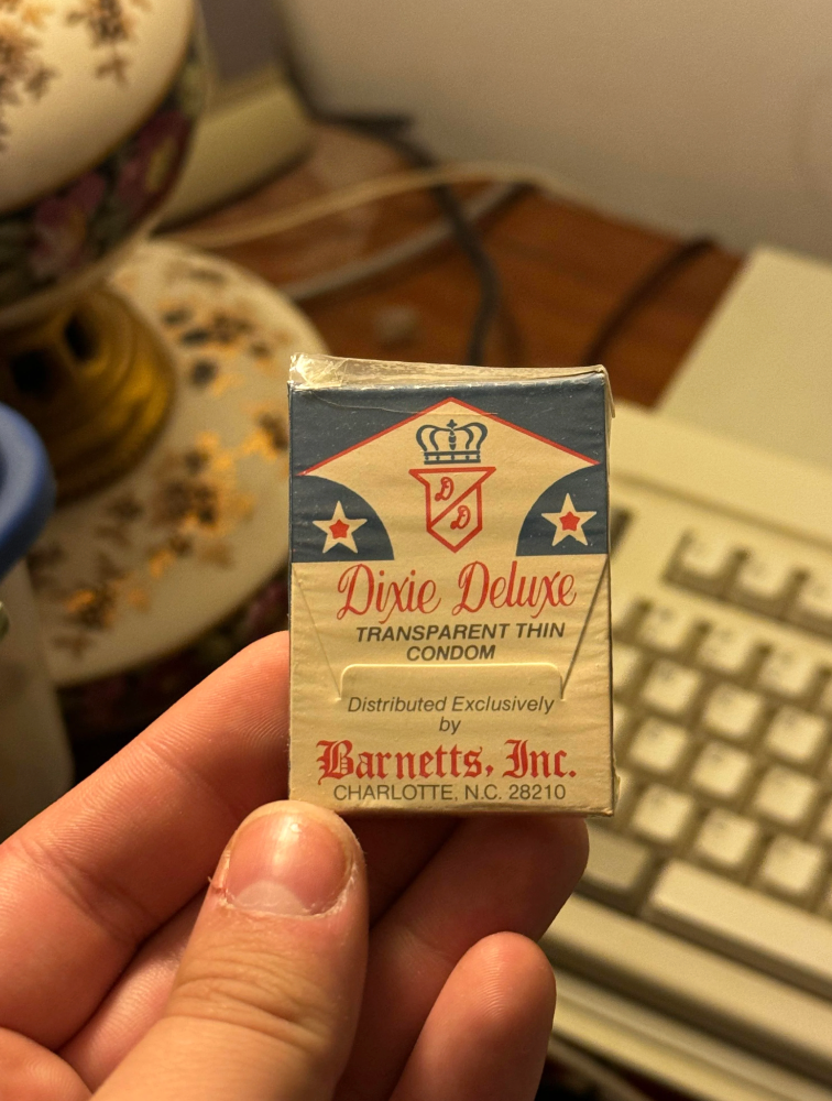 A person's hand holds a vintage "Dixie Deluxe" condom package with a retro design, featuring the text "Transparent Thin Condom."