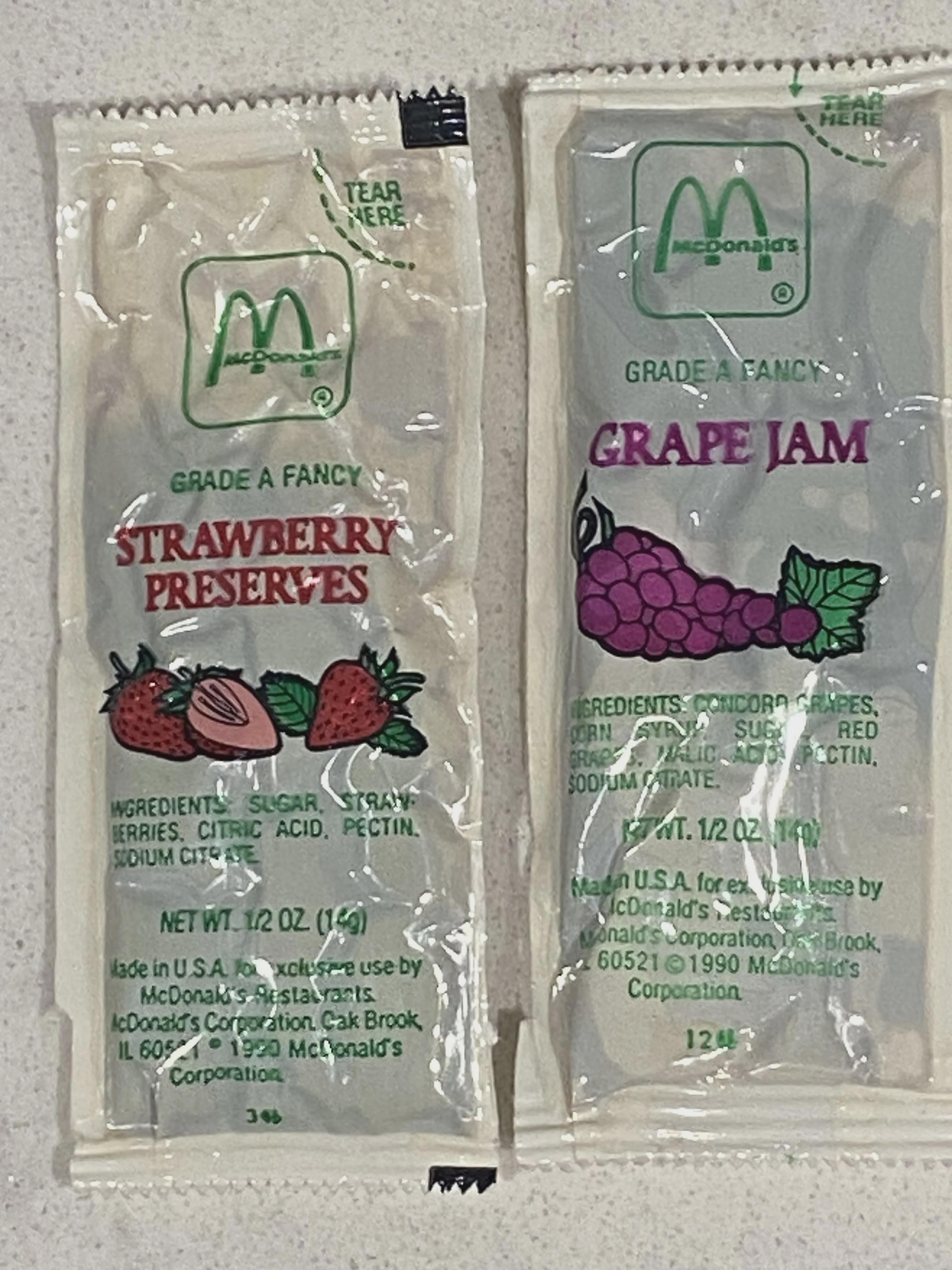 Two McDonald's jam packets: one strawberry preserves with an illustration of strawberries, and the other grape jam with an illustration of grapes