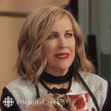 A character from "Schitt's Creek" sips from a floral mug, looking surprised. Text overlay: "#SchittsCreek."