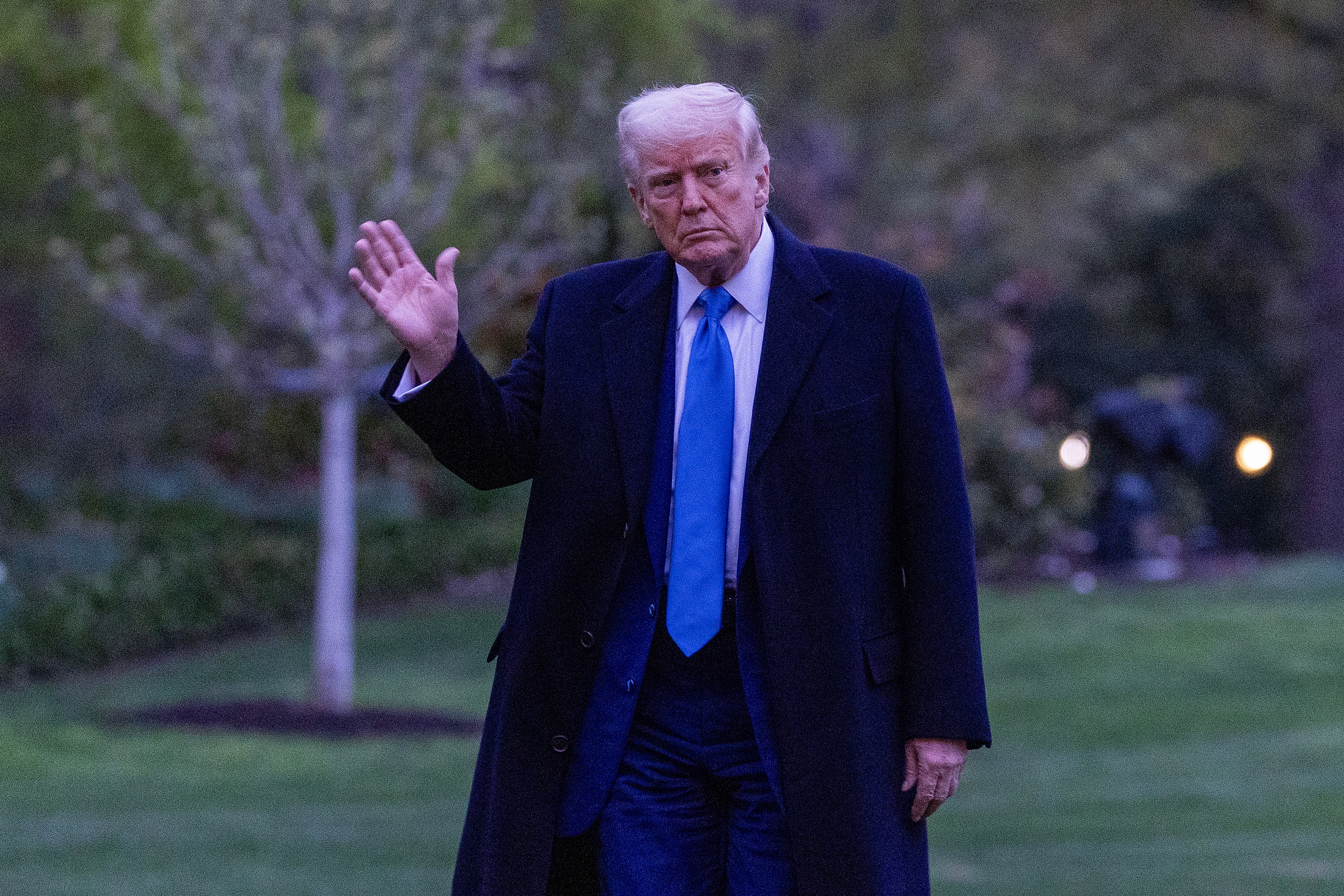A man in a suit and long coat waves while walking outdoors, appearing serious. Trees and a lawn are in the background