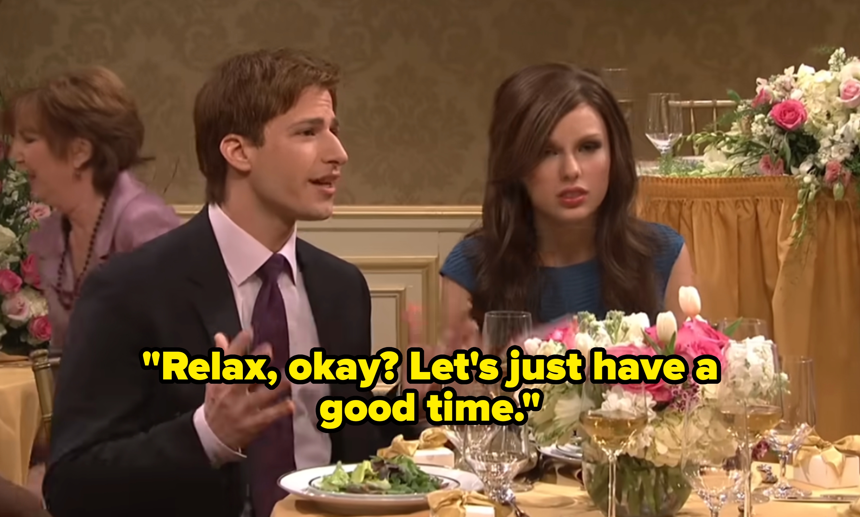A man and woman sit at a wedding reception table, engaged in conversation, surrounded by floral centerpieces