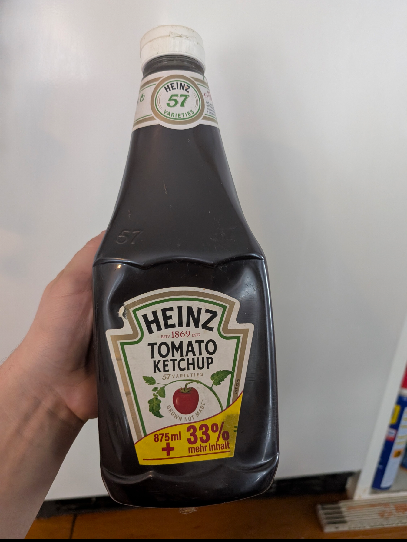 A hand holds a bottle of Heinz Tomato Ketchup featuring German text and a 33% more content label