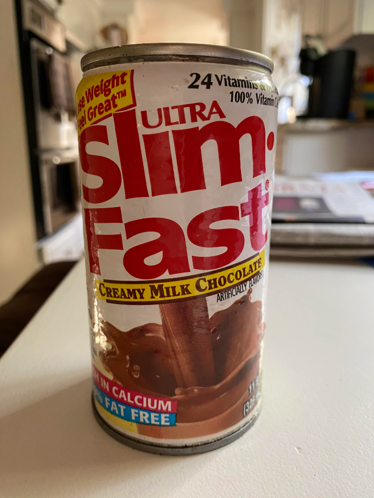 SlimFast can on a table with "Ultra SlimFast", "Creamy Milk Chocolate", "24 Vitamins & Minerals", "0% Fat" and "All Essential Nutrients" text