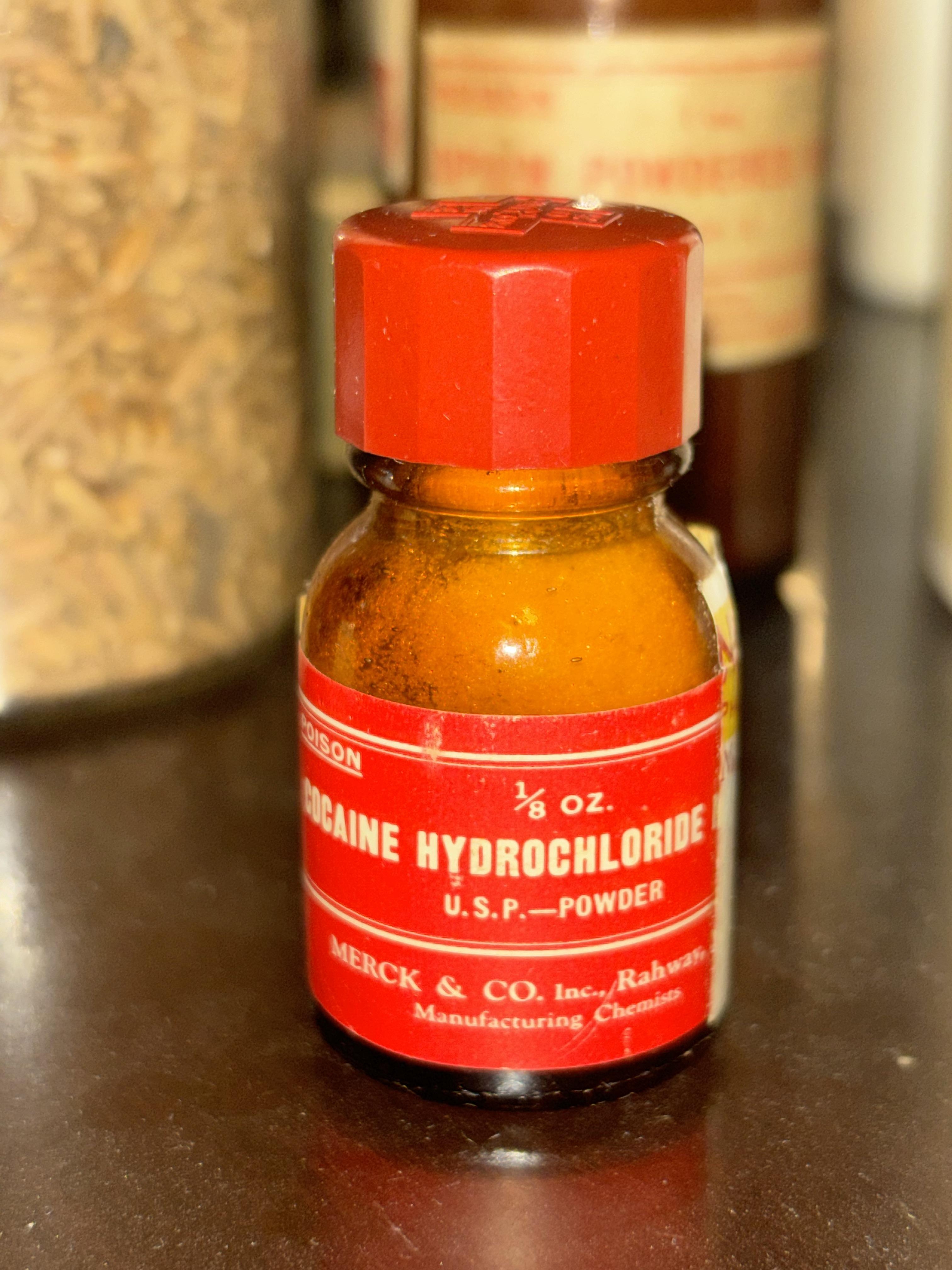 Small vintage bottle labeled "Cocaine Hydrochloride, U.S.P. - Powder," manufactured by Merck & Co. Bottle has a red cap and label