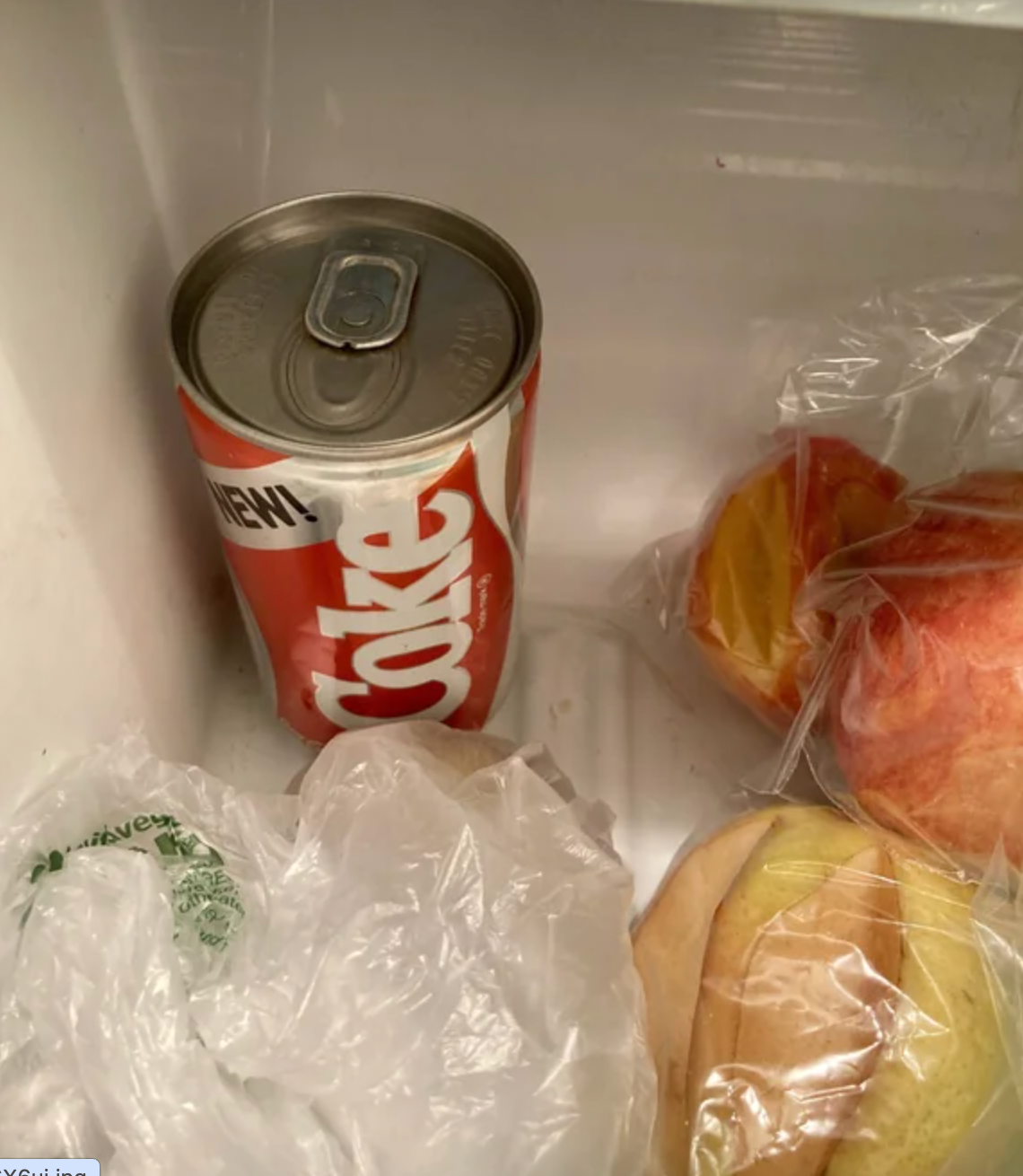 Soda can in fridge beside bagged fruit, including oranges and apples