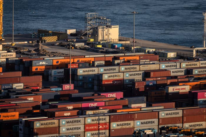 Stacks of shipping containers at a busy port near the water, with cranes and industrial buildings in the background