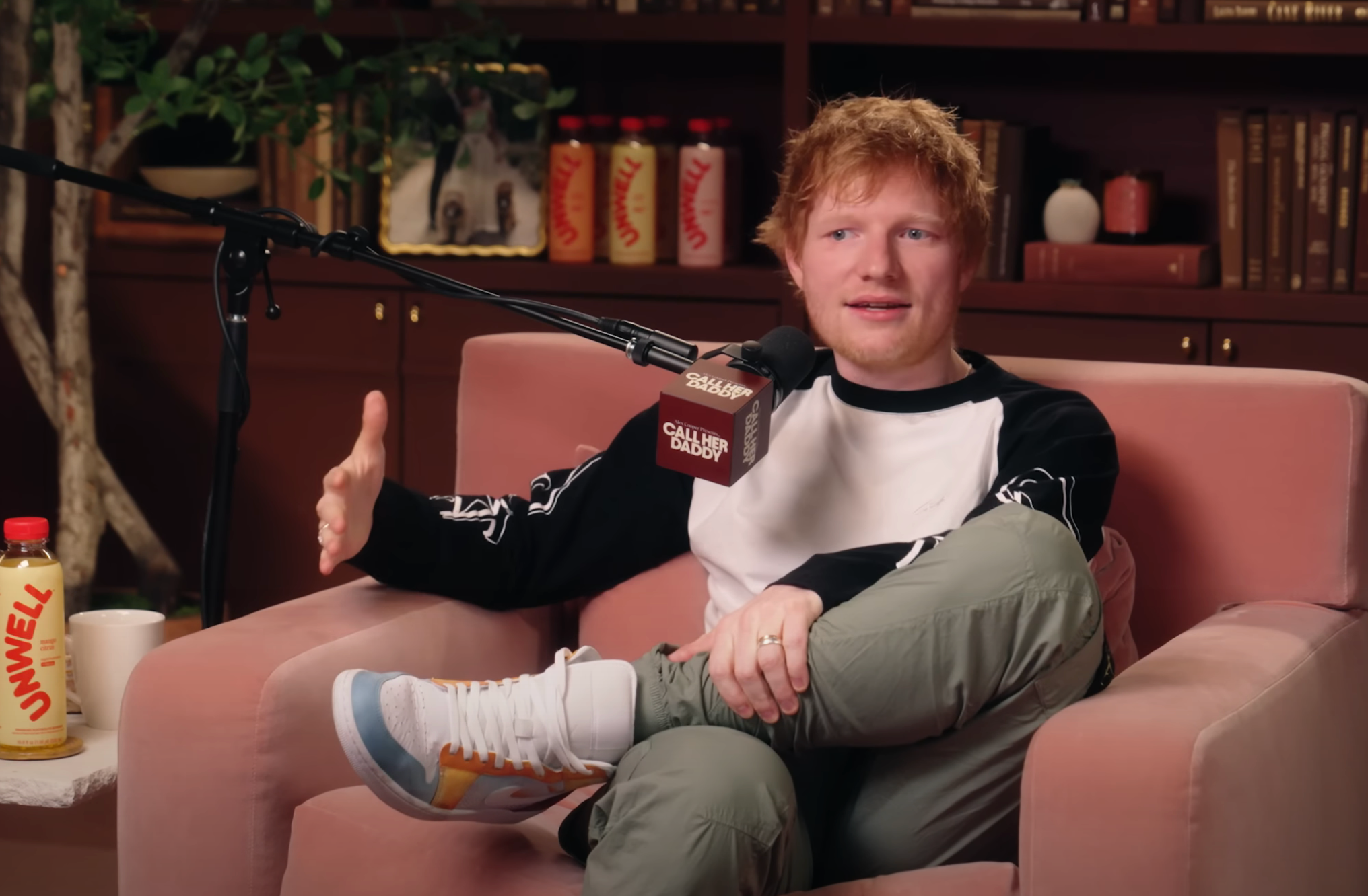 Person in a casual setting, wearing a long-sleeve shirt and sneakers, speaking into a microphone during an interview