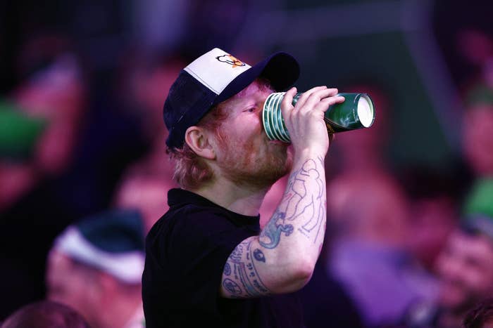 A person wearing a hat drinks from stacked cups at a public event, with tattooed arm visible