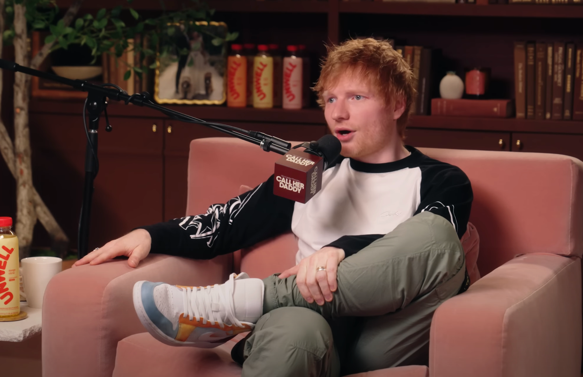 Musician in casual wear, seated on a pink couch, speaking into a microphone during a podcast interview