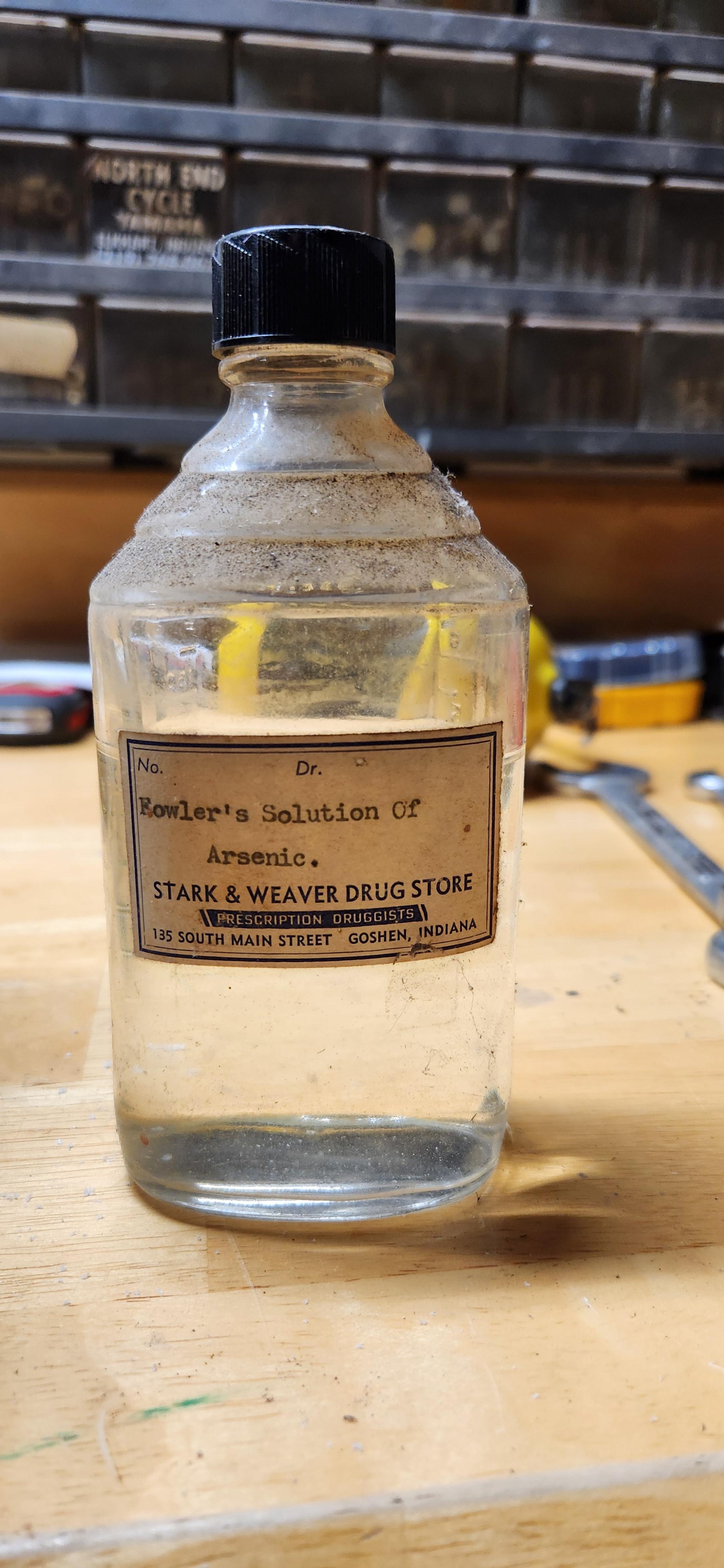 Old medicinal bottle labeled "Fowler's Solution of Arsenic," from Stark & Weaver Drug Store, Goshen, Indiana, on a cluttered workbench
