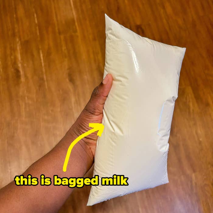A hand holds a sealed plastic bag filled with liquid, likely milk, against a wooden floor background