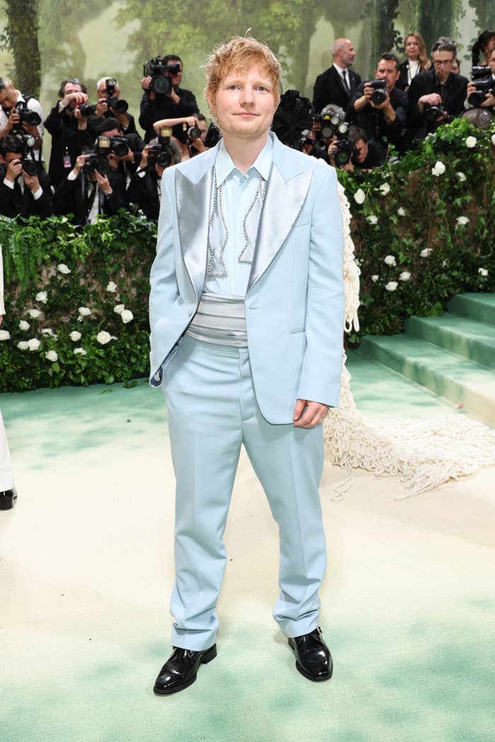Man in light-colored suit with satin lapels, standing on an event carpet surrounded by photographers