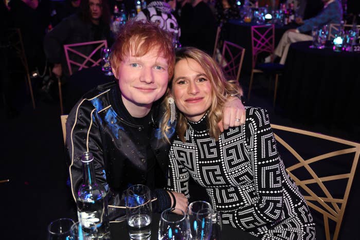 Two people smiling at an event. The person on the left wears a dark, decorated jacket; the person on the right, a patterned black-and-white outfit