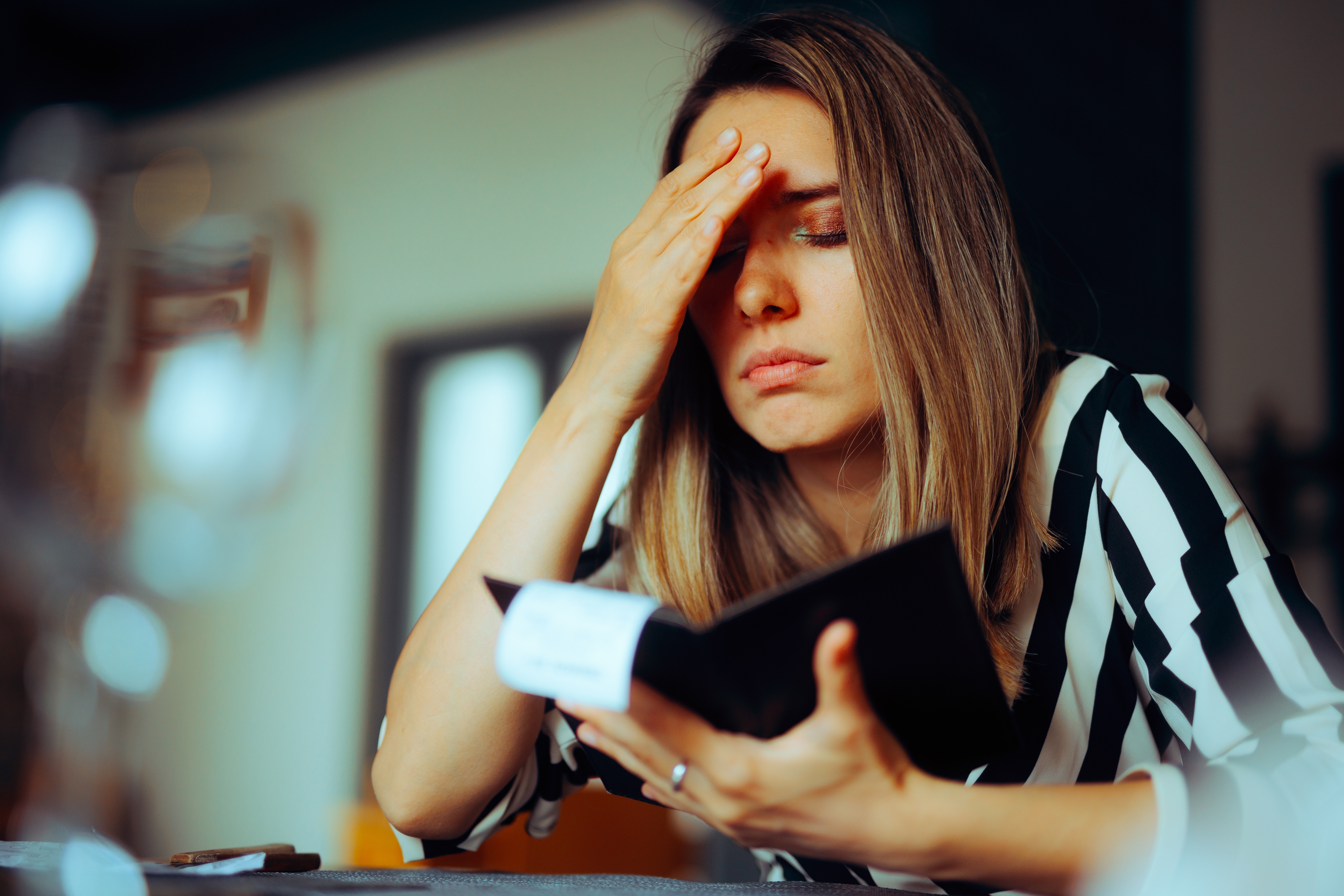 A woman in a striped shirt holds her forehead in frustration while looking at a bill inside a wallet