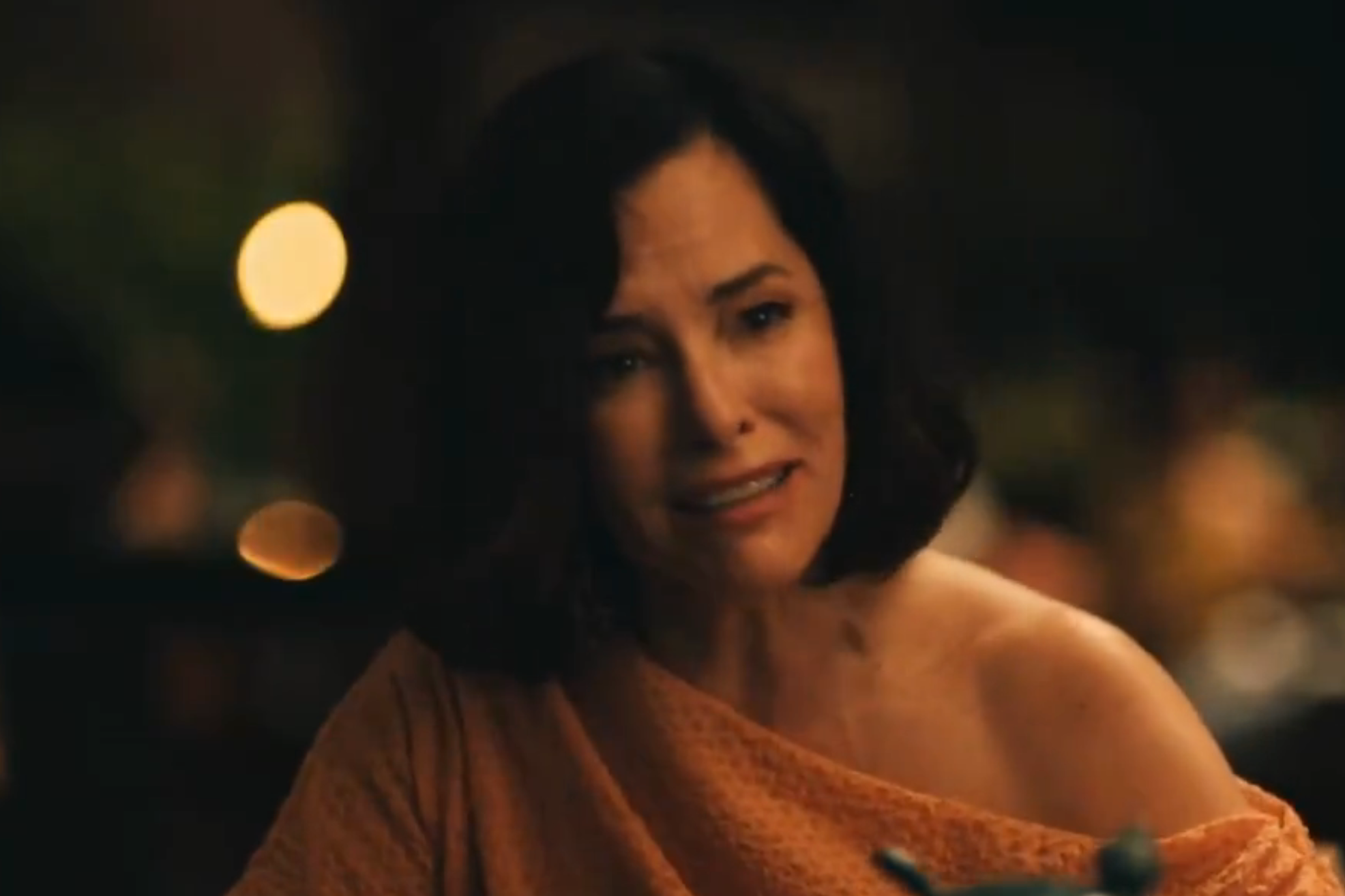 A person with short dark hair wearing an off-the-shoulder top looks emotional in a dimly lit setting