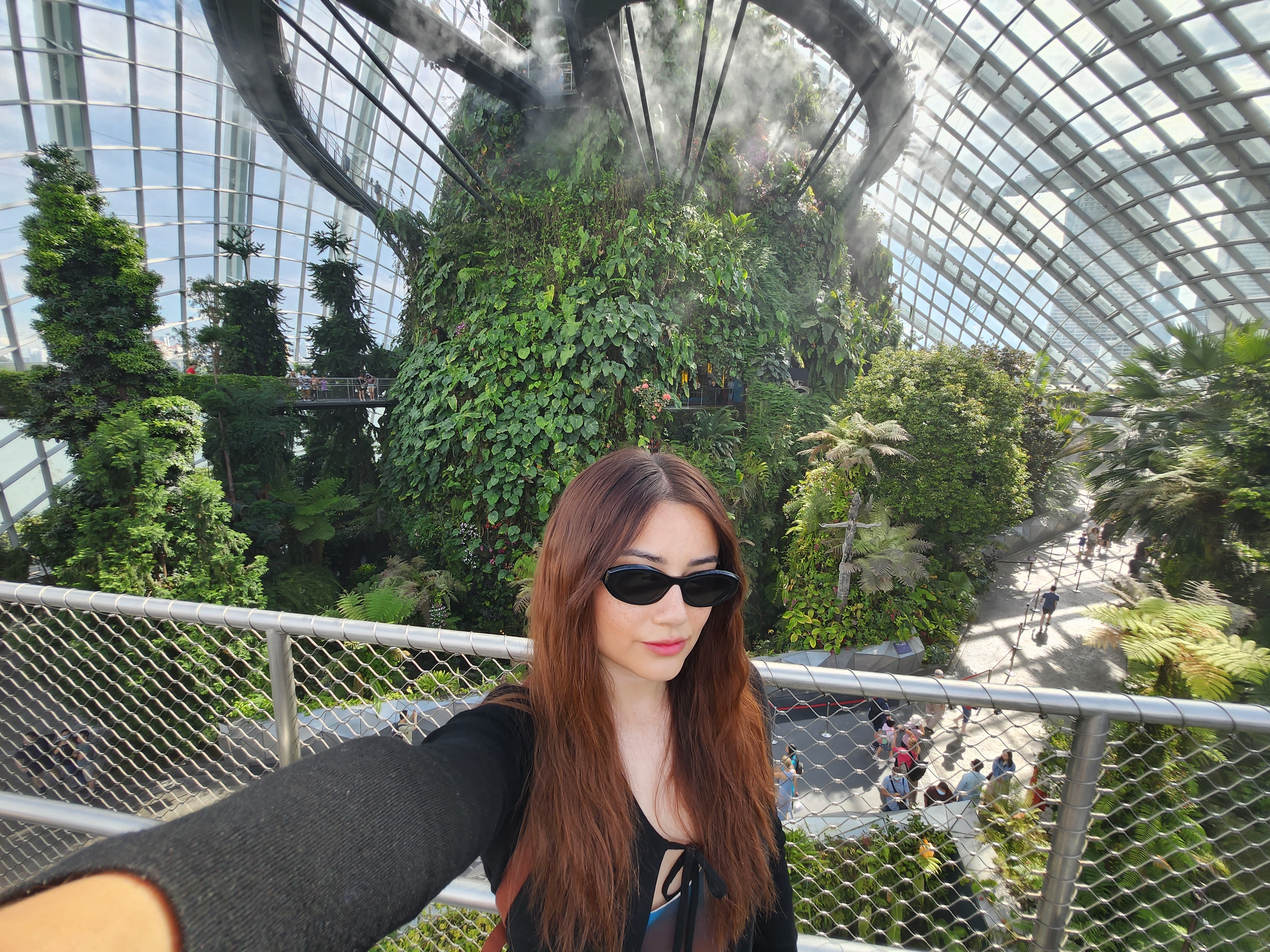 The author, with long hair and sunglasses, takes a selfie in a lush, greenhouse-like conservatory with tropical plants and a high glass ceiling