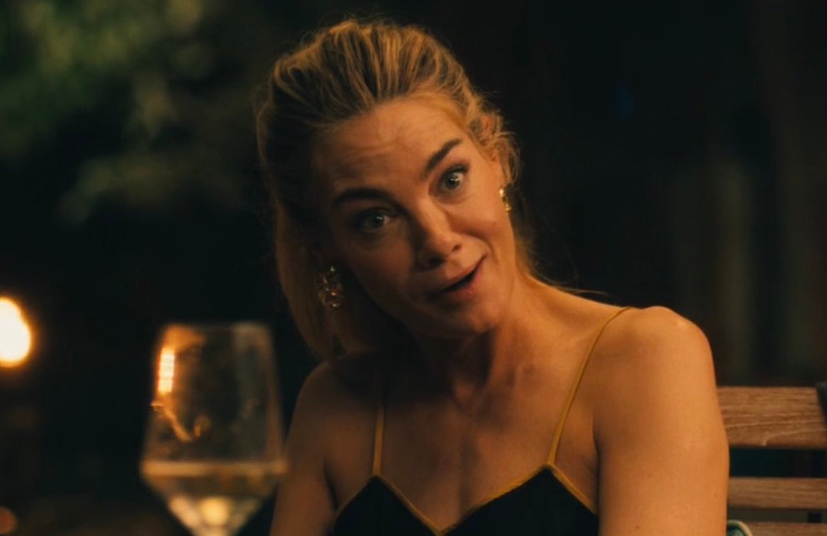 Woman at an outdoor dinner table expresses surprise, holding a glass. Elegant dress and earrings suggest a social gathering