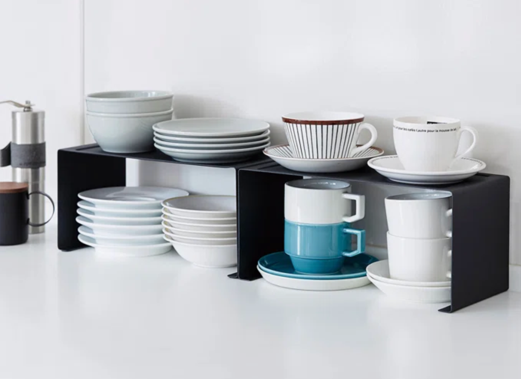 Black countertop organizer holding neatly stacked bowls, plates, and mugs. Kitchenware is arranged efficiently on two levels for space-saving