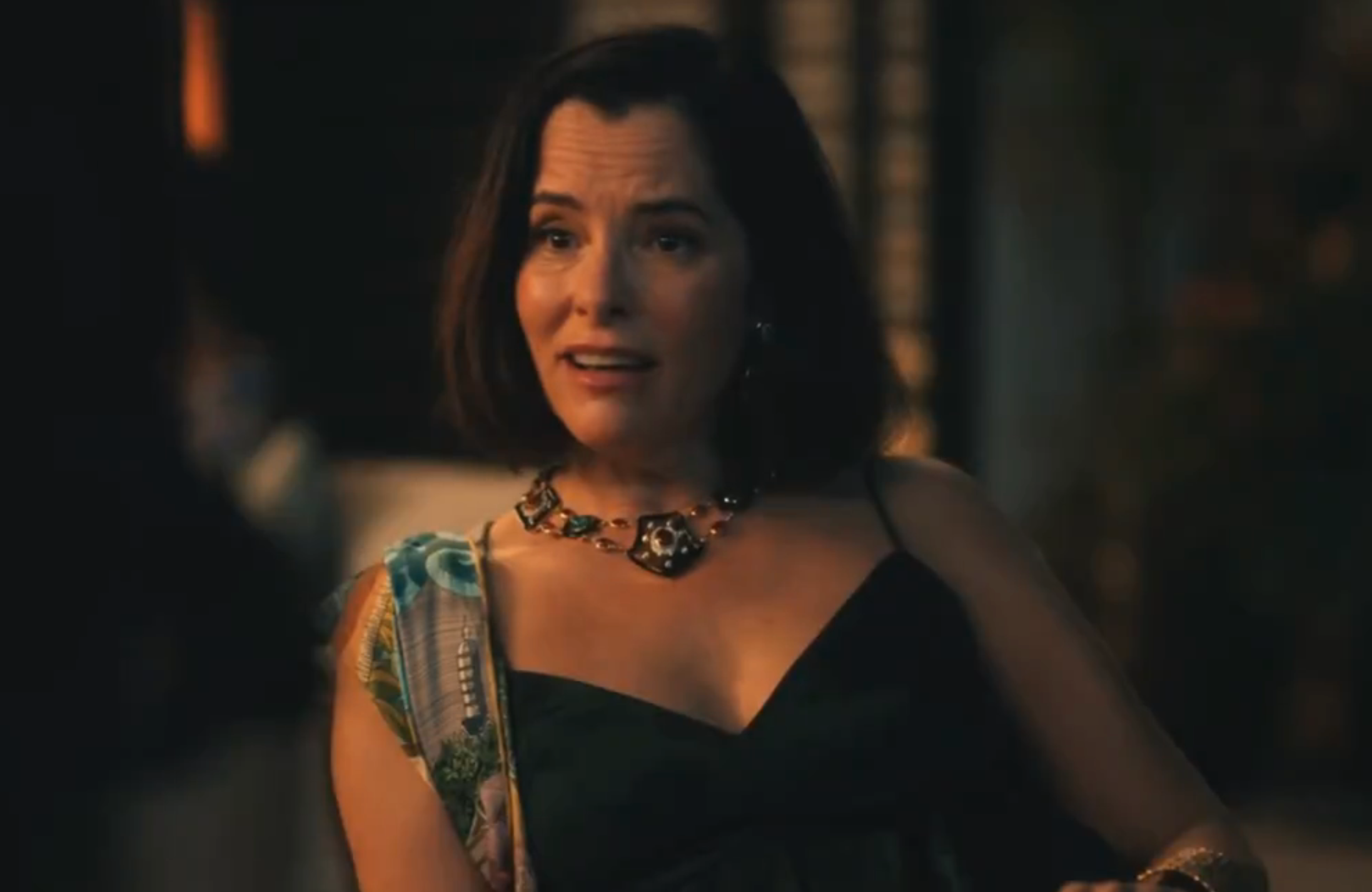 Person in elegant evening attire, featuring a V-neck dress and statement necklace, sitting and engaged in a conversation