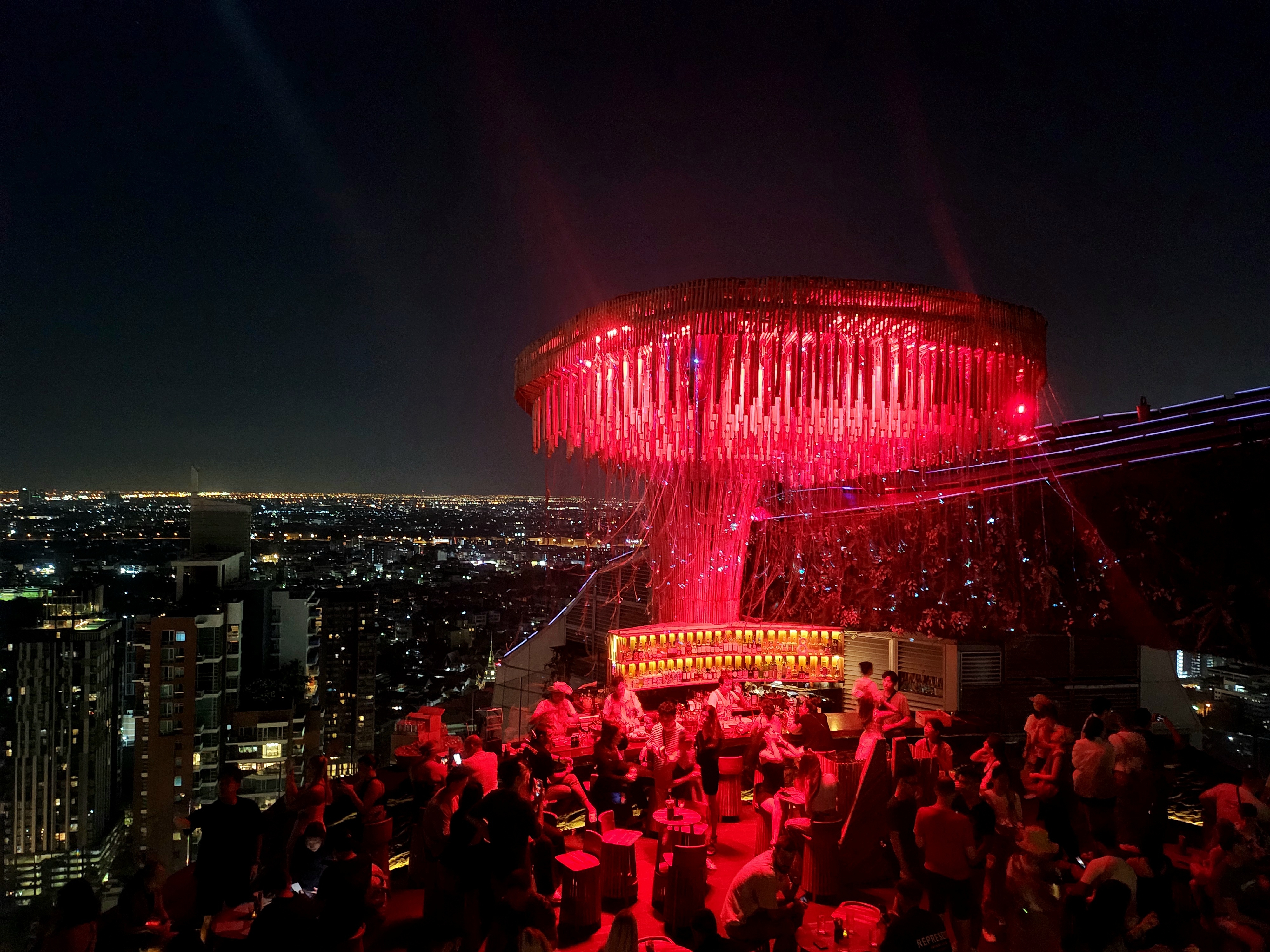 Rooftop bar with a large, illuminated chandelier overlooking a cityscape at night. Crowded with people socializing and taking in the view