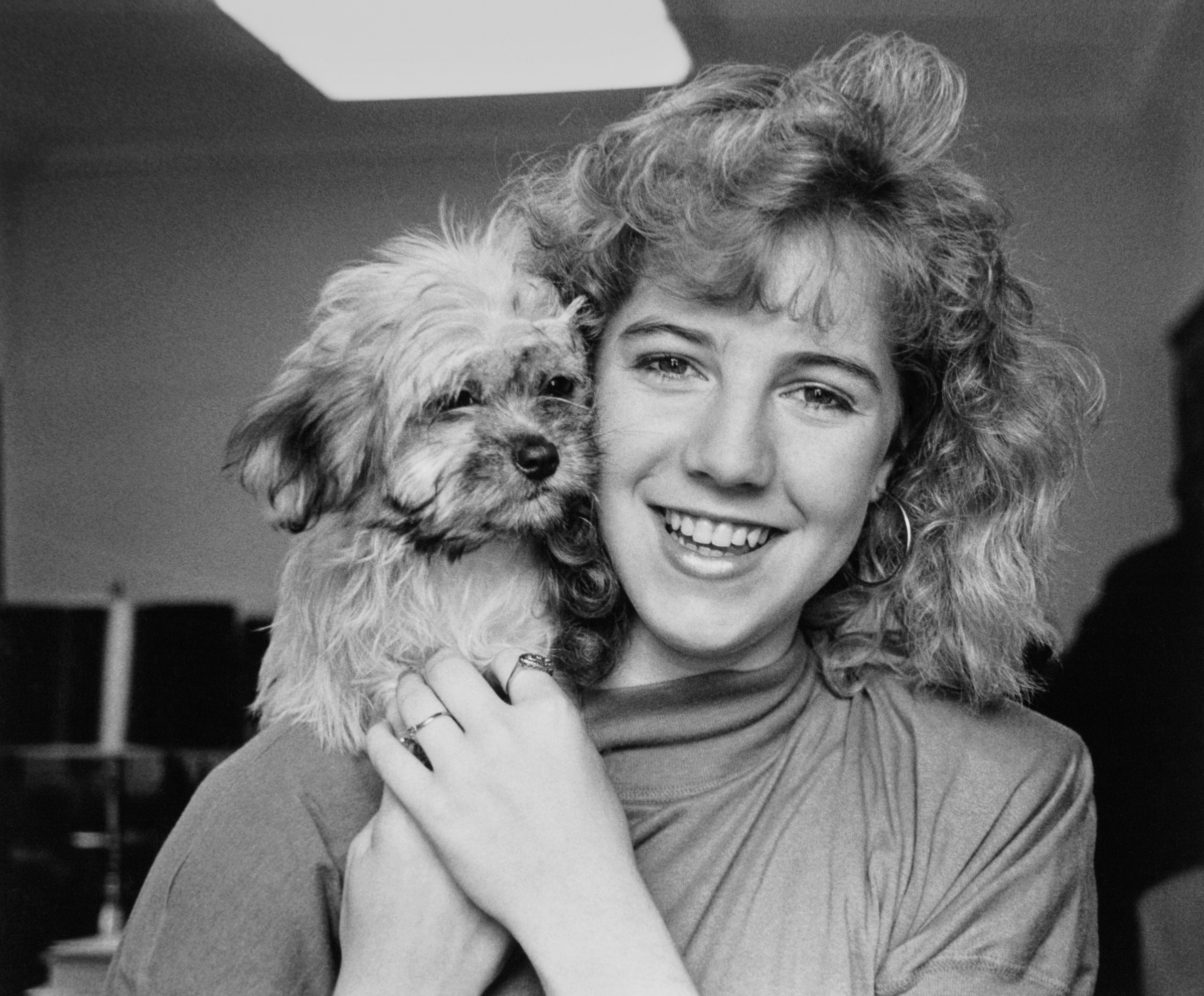 Person with curly hair smiling and holding a small, fluffy dog close to their face indoors