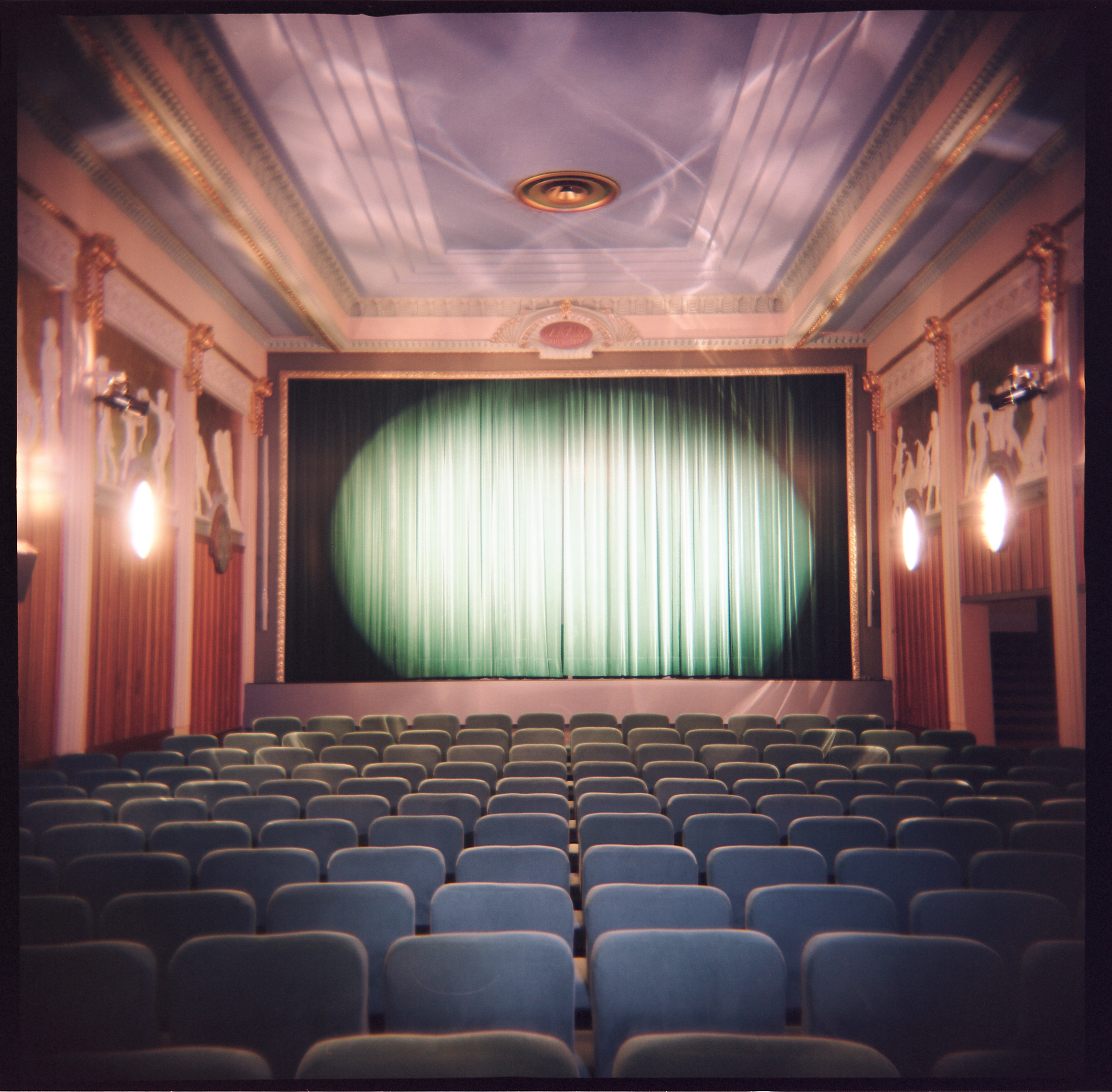 Vintage theater interior with ornate ceiling, decorative wall panels, and rows of empty seats facing a closed stage curtain