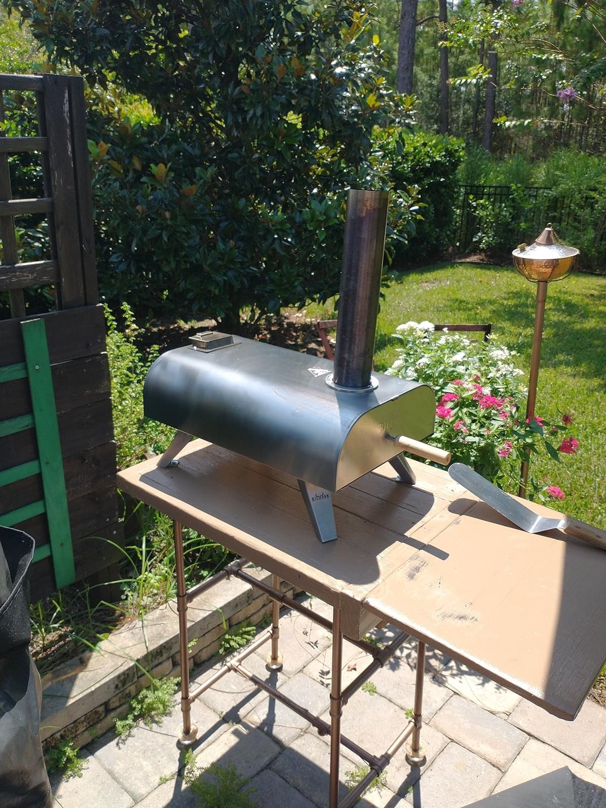 Portable outdoor pizza oven connected  a table, surrounded by a plot  with flowers and greenery, nether  a sunny sky