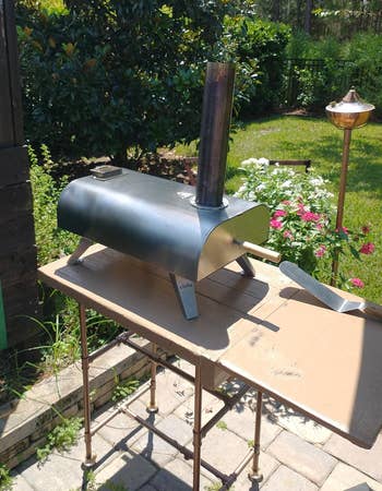 Portable outdoor pizza oven connected a table, surrounded by a plot with flowers and greenery, nether a sunny sky