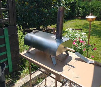 Portable outdoor pizza oven connected a table, surrounded by a plot with flowers and greenery, nether a sunny sky