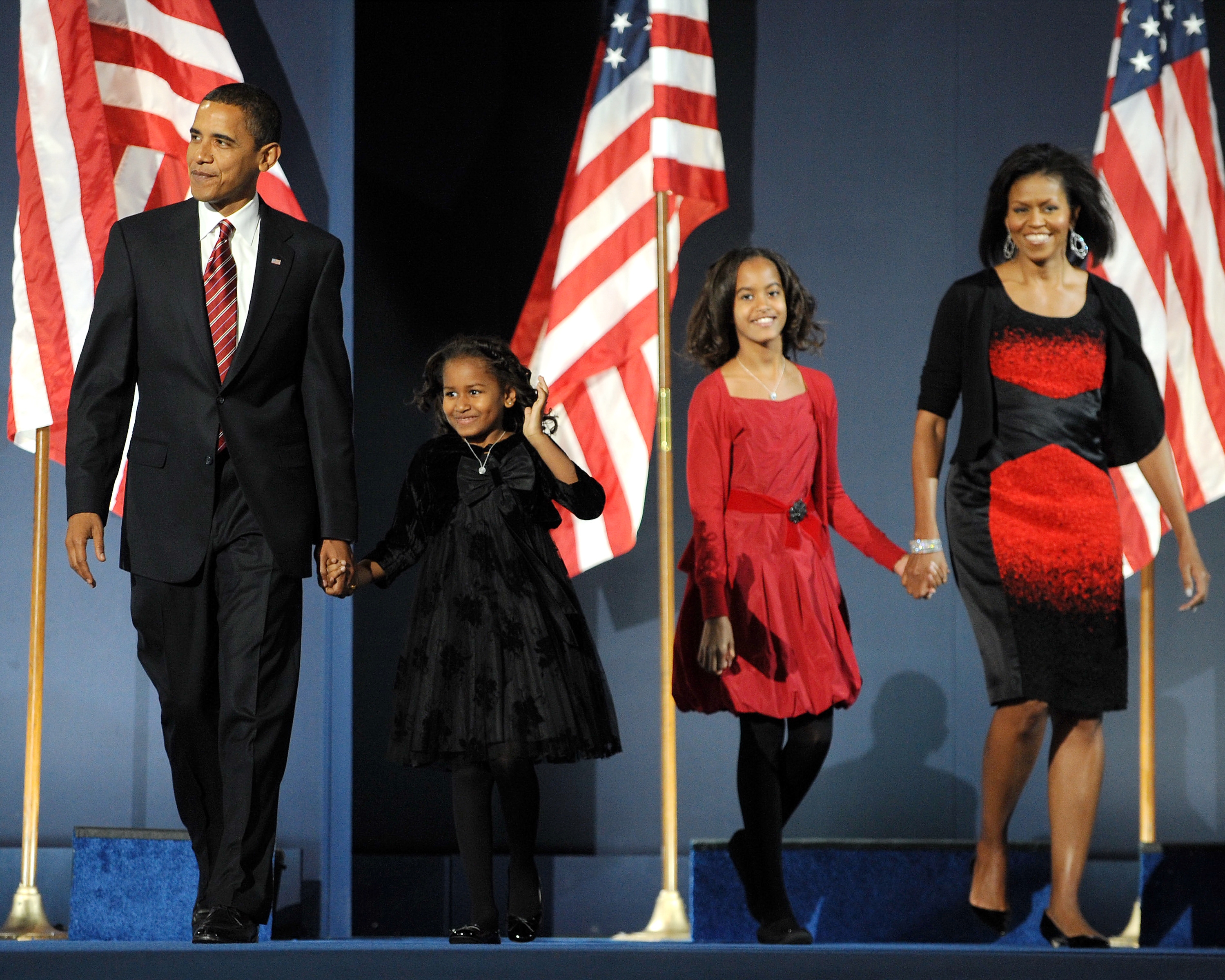 A family walks hand in hand on stage with U.S. flags in the background. The woman wears a stylish, fitted dress, the others are in formal attire