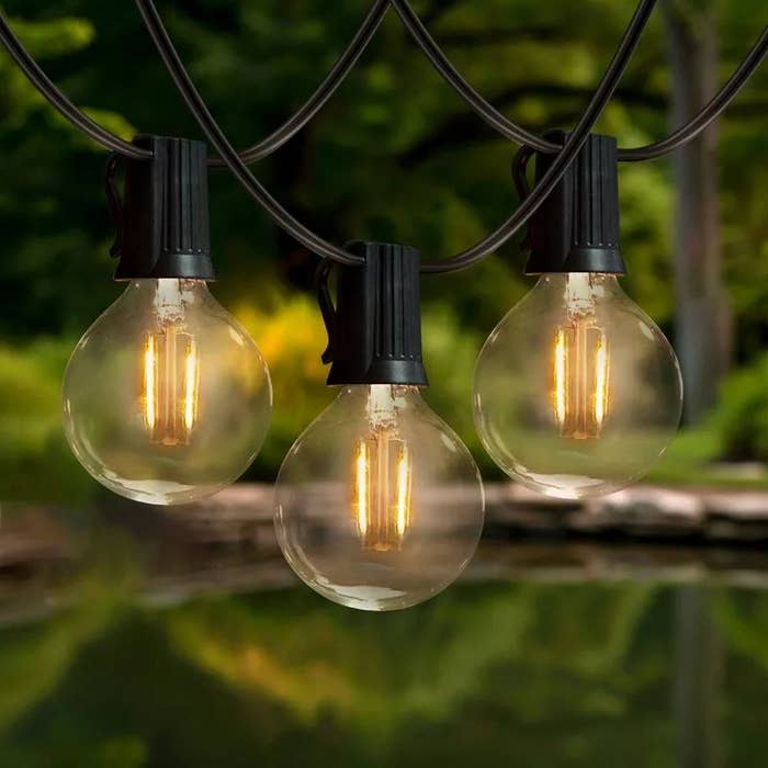 Three decorative outdoor string lights with visible filaments hang in front of a serene, blurred natural background. Ideal for garden ambiance
