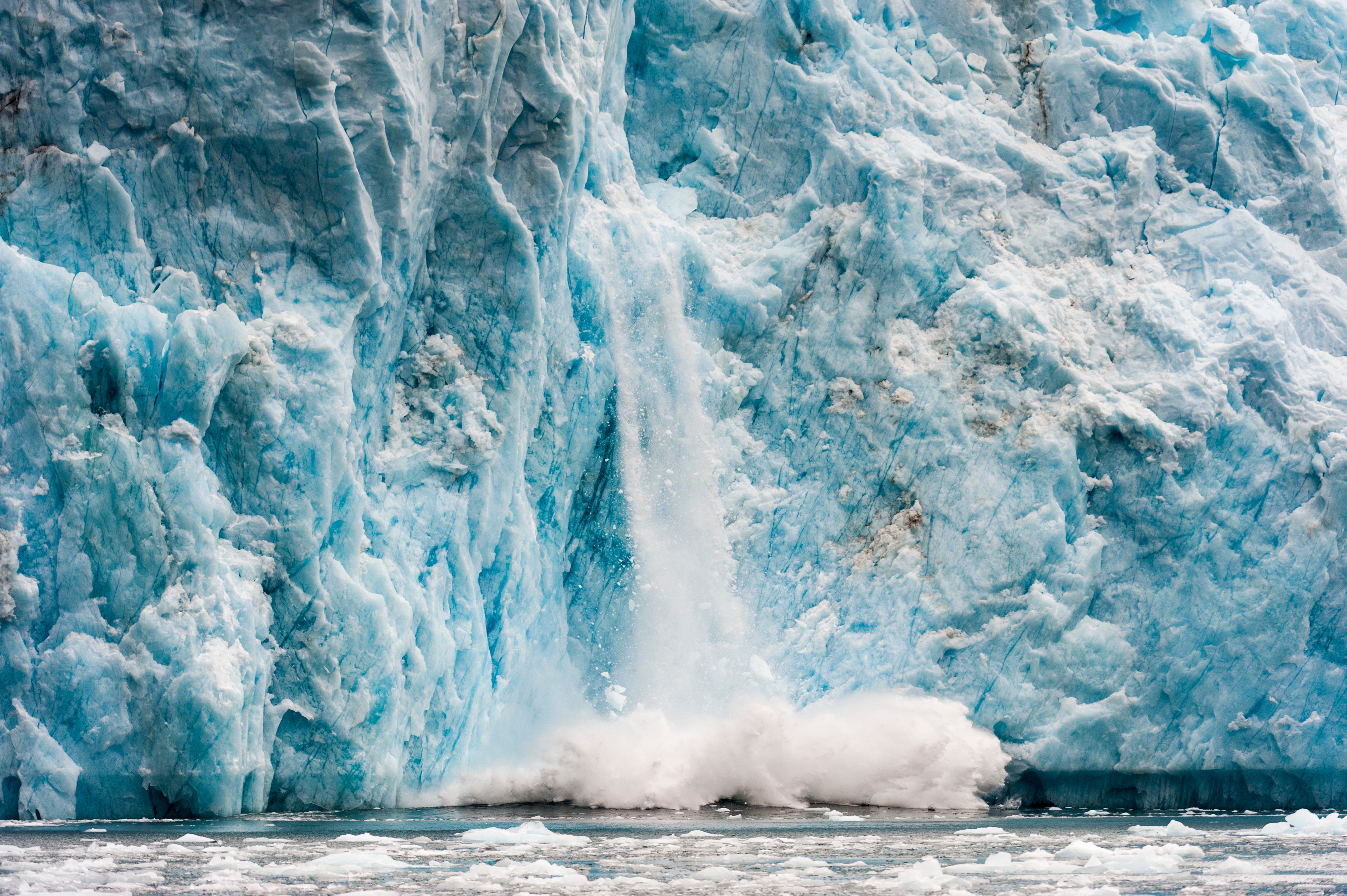 A waterfall flows down a massive glacier, crashing into icy waters below, creating mist and splashes