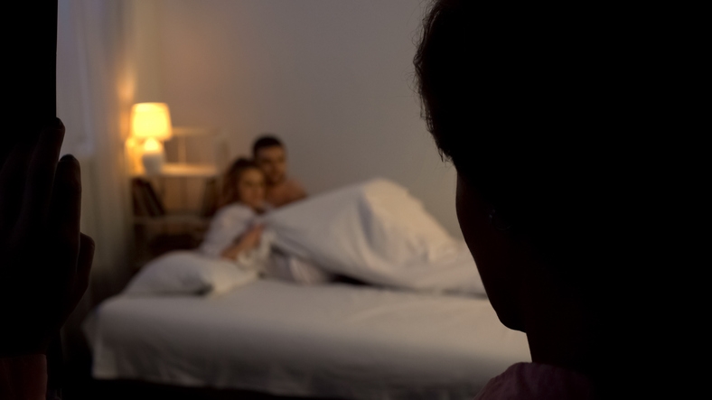 Two people in bed, partially covered by a sheet, seem surprised as someone watches from the doorway