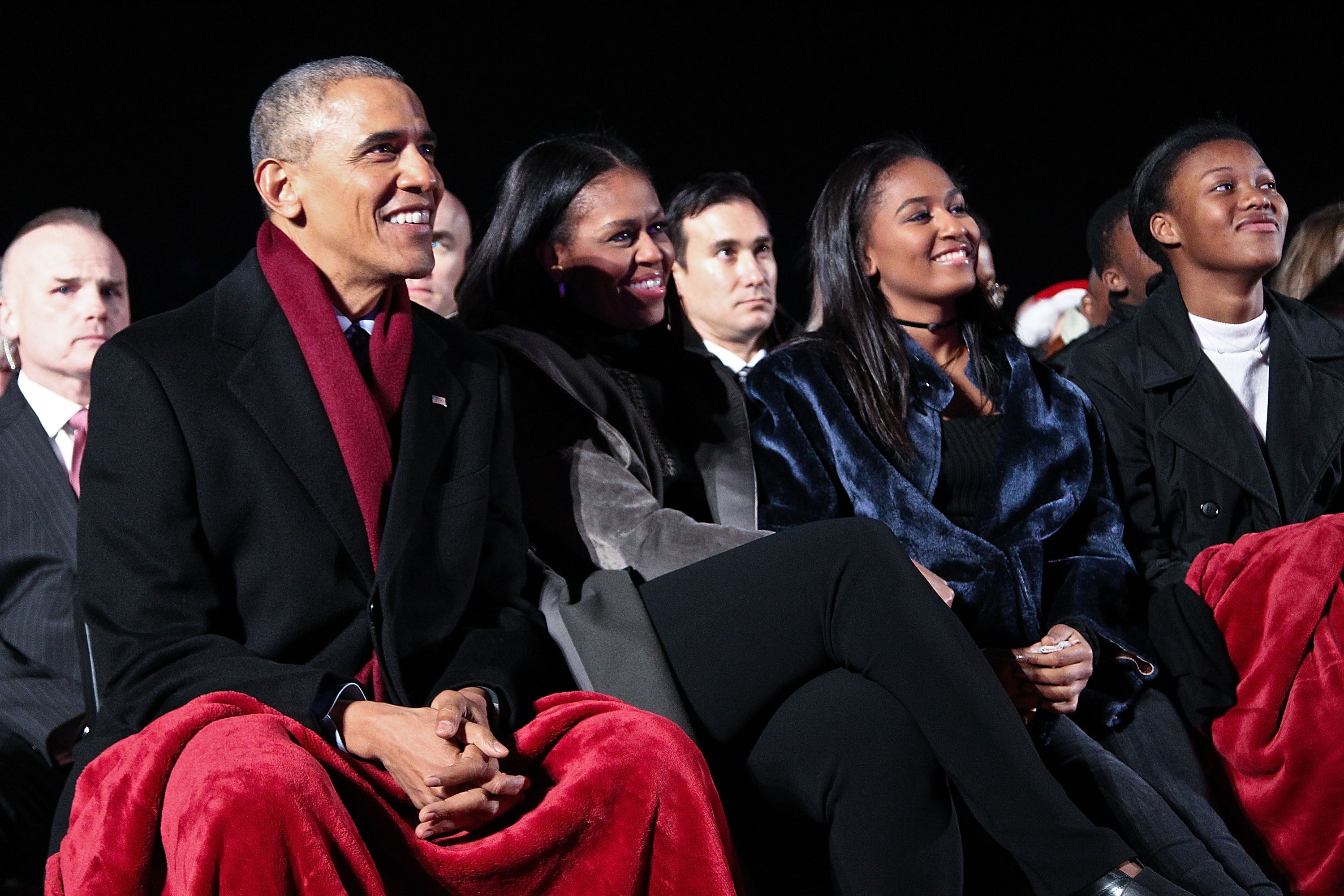 A group of four people, including a well-dressed family, sitting together at an event, wrapped in blankets, looking ahead and smiling