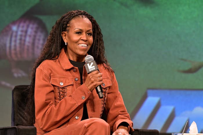 A woman smiles on stage, holding a microphone labeled "SXSW." She is wearing a casual brown jacket and has long braids