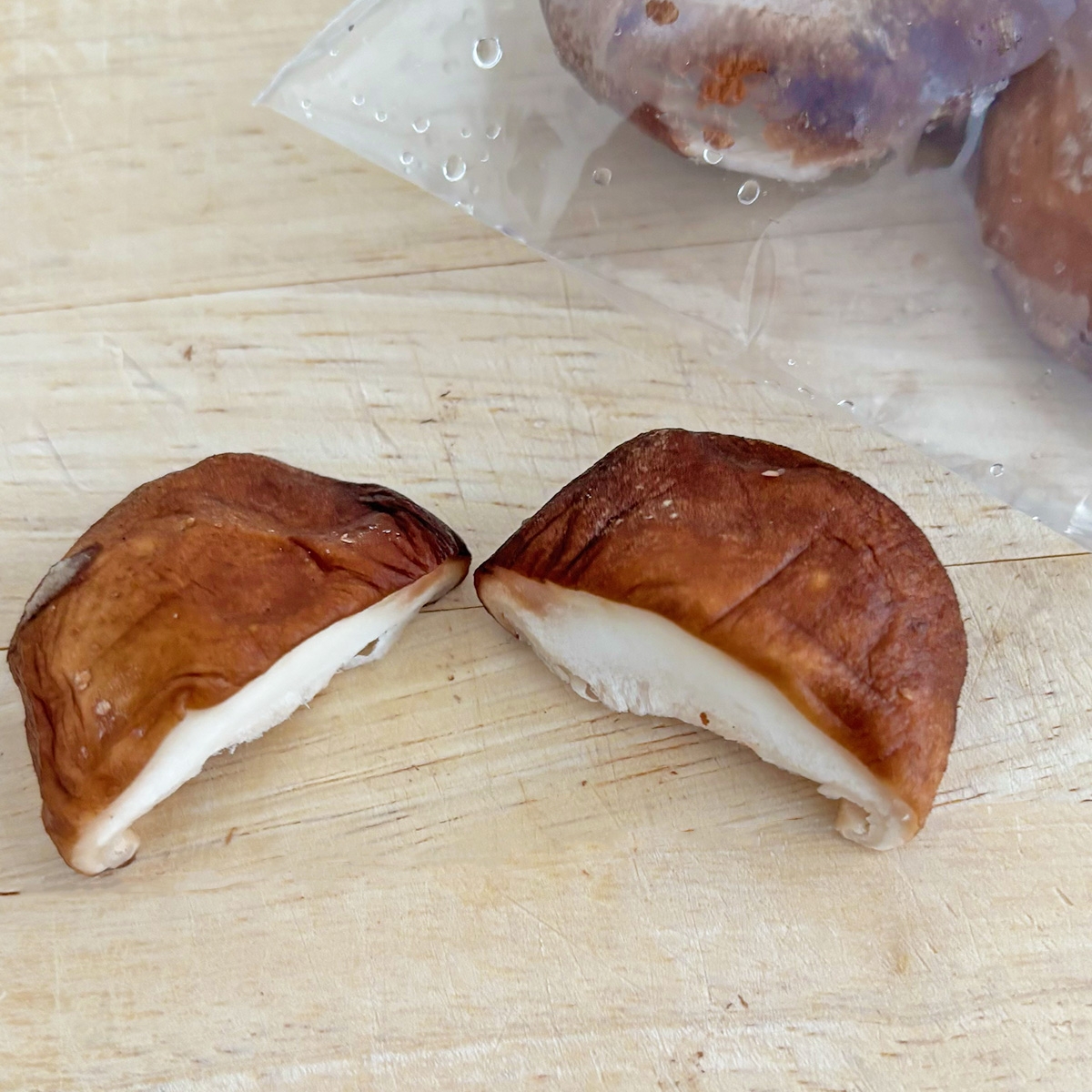 椎茸の半分に切られた部分がまな板の上に置かれている。後ろに袋入りの椎茸も見える。